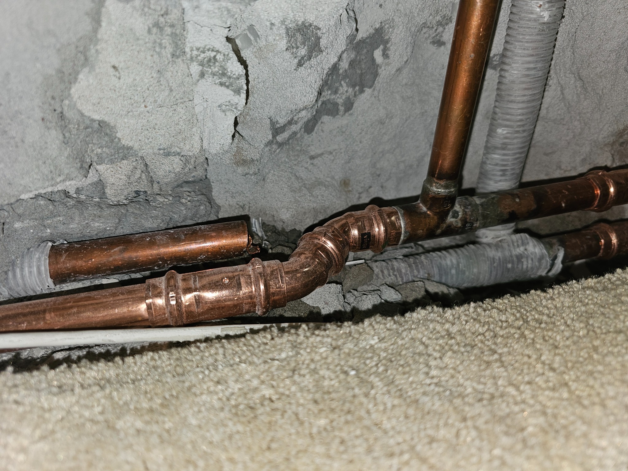 Miedziane rury instalacji centralnego ogrzewania połączone metodą zaprasowywania, zamontowane przy szarej, nierównej ścianie. Widoczna również rura karbowana. Część instalacji ukryta za wykładziną...