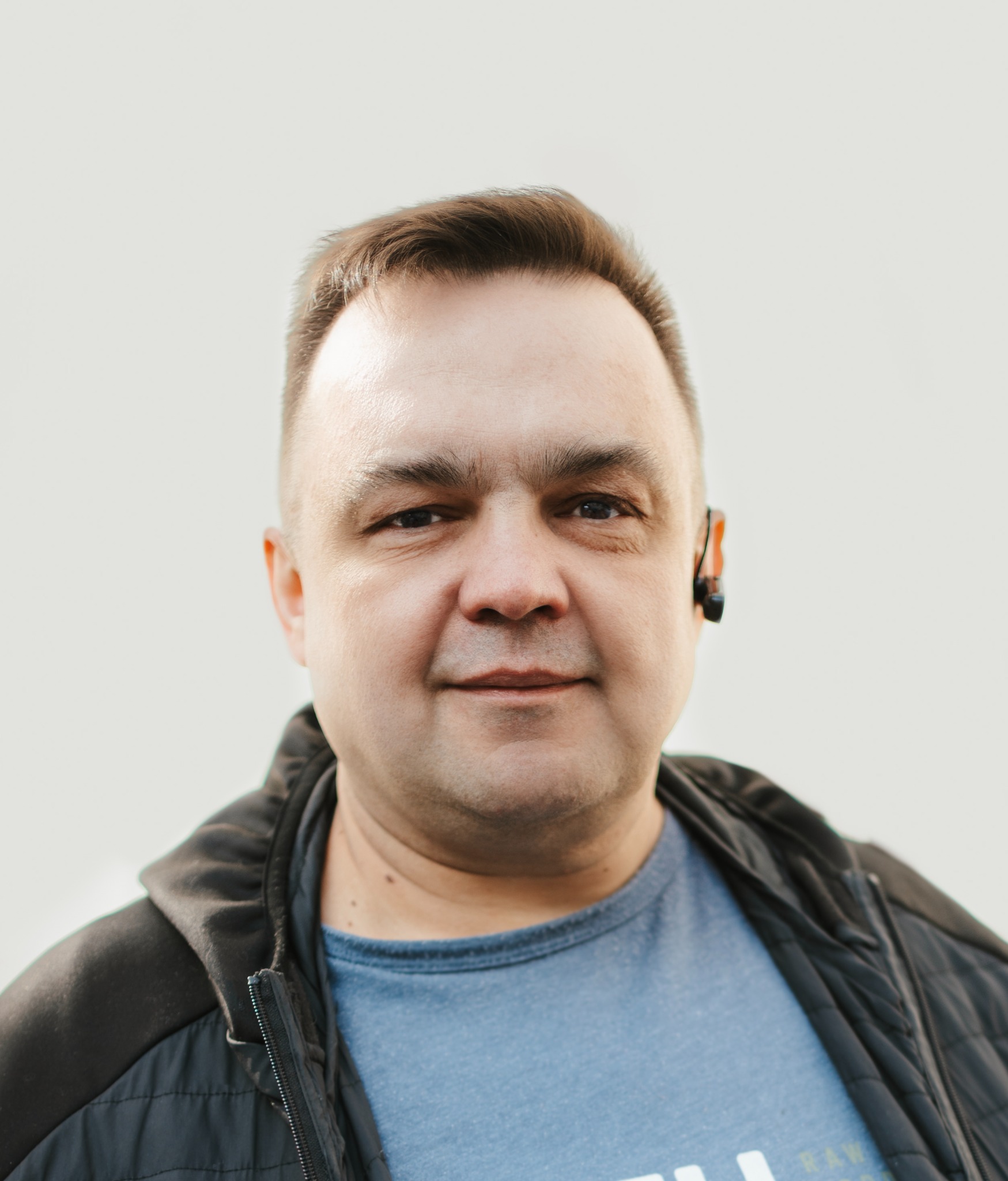 Portret mężczyzny z zestawem słuchawkowym Bluetooth, ubrany w niebieską koszulkę i czarną kurtkę, na jasnym tle.