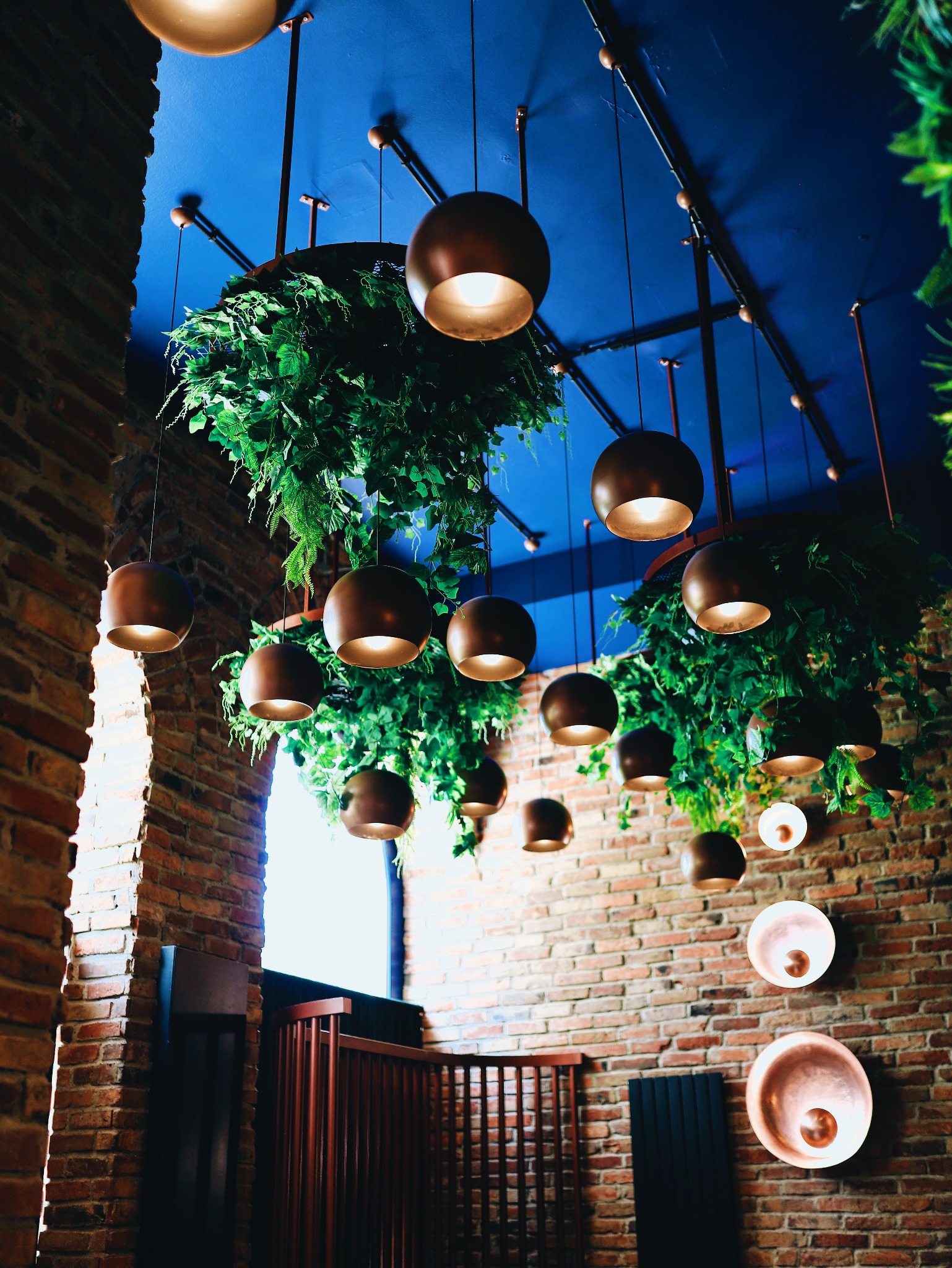Industrialna aranżacja wnętrza z ceglaną ścianą, wiszącymi kulistymi lampami i zielonymi roślinami, tworząca nastrojowe oświetlenie.