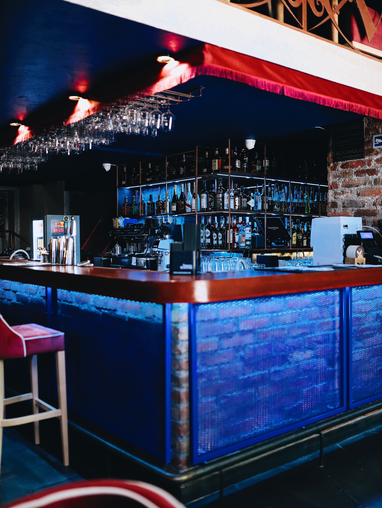 Nowoczesny bar z drewnianym blatem, podświetlonym na niebiesko panelem z siatki i ceglanymi detalami; w tle półki z butelkami i wiszące kieliszki.
