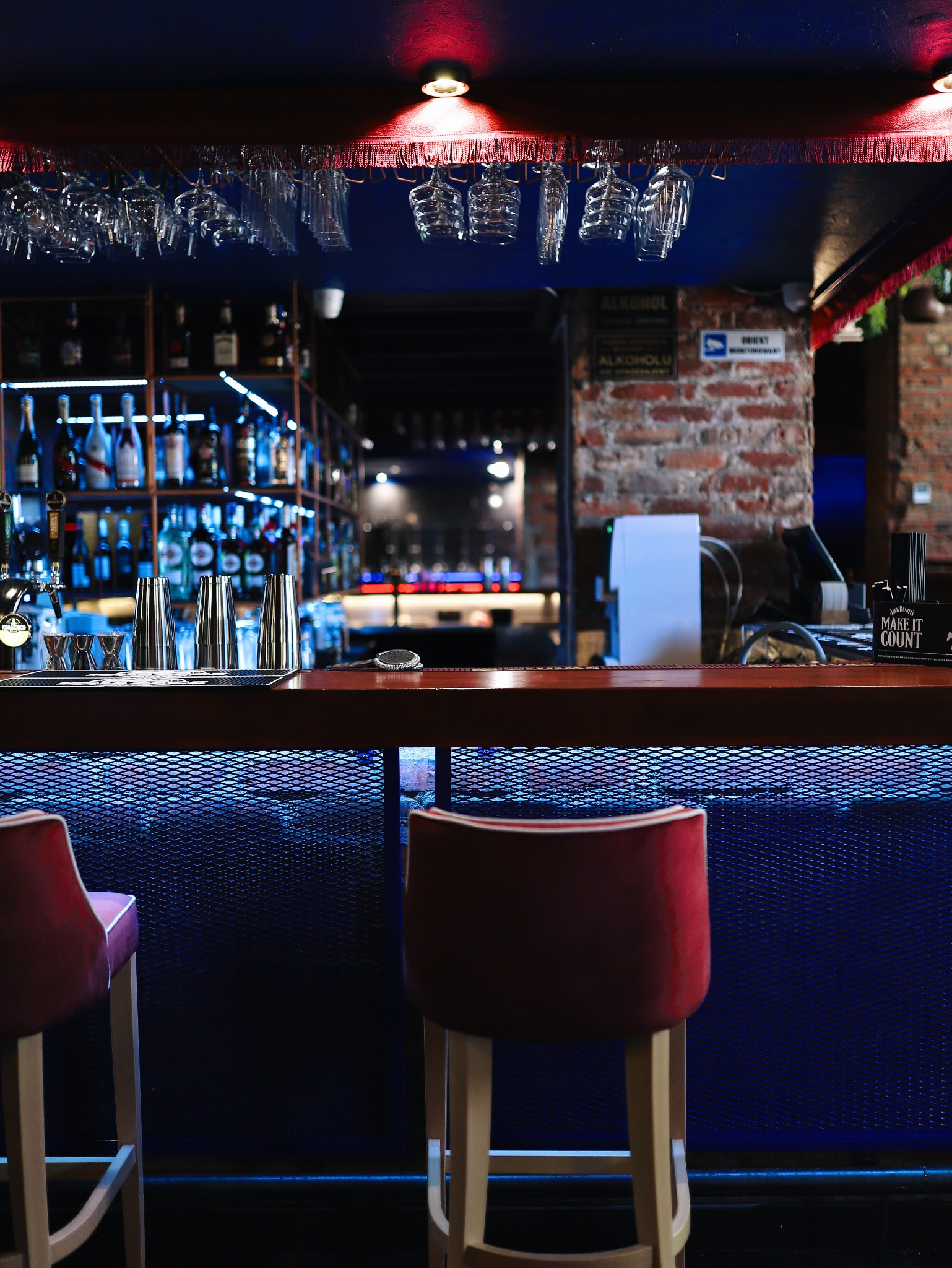 Profesjonalnie zaaranżowany bar z rzędami butelek alkoholu na półkach, metalowymi shakerami i eleganckimi kieliszkami wiszącymi nad blatem, oświetlony niebieskim światłem, z dwoma czerwonymi...