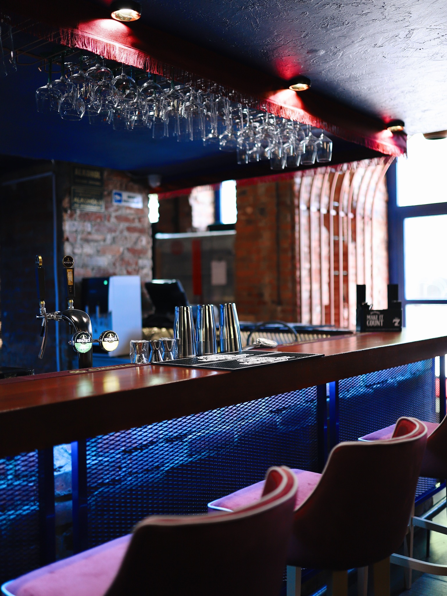 Nowoczesny bar z drewnianym blatem, podświetlany na niebiesko, z wiszącymi kieliszkami i różowymi krzesłami barowymi, widok na ceglaną ścianę w tle.