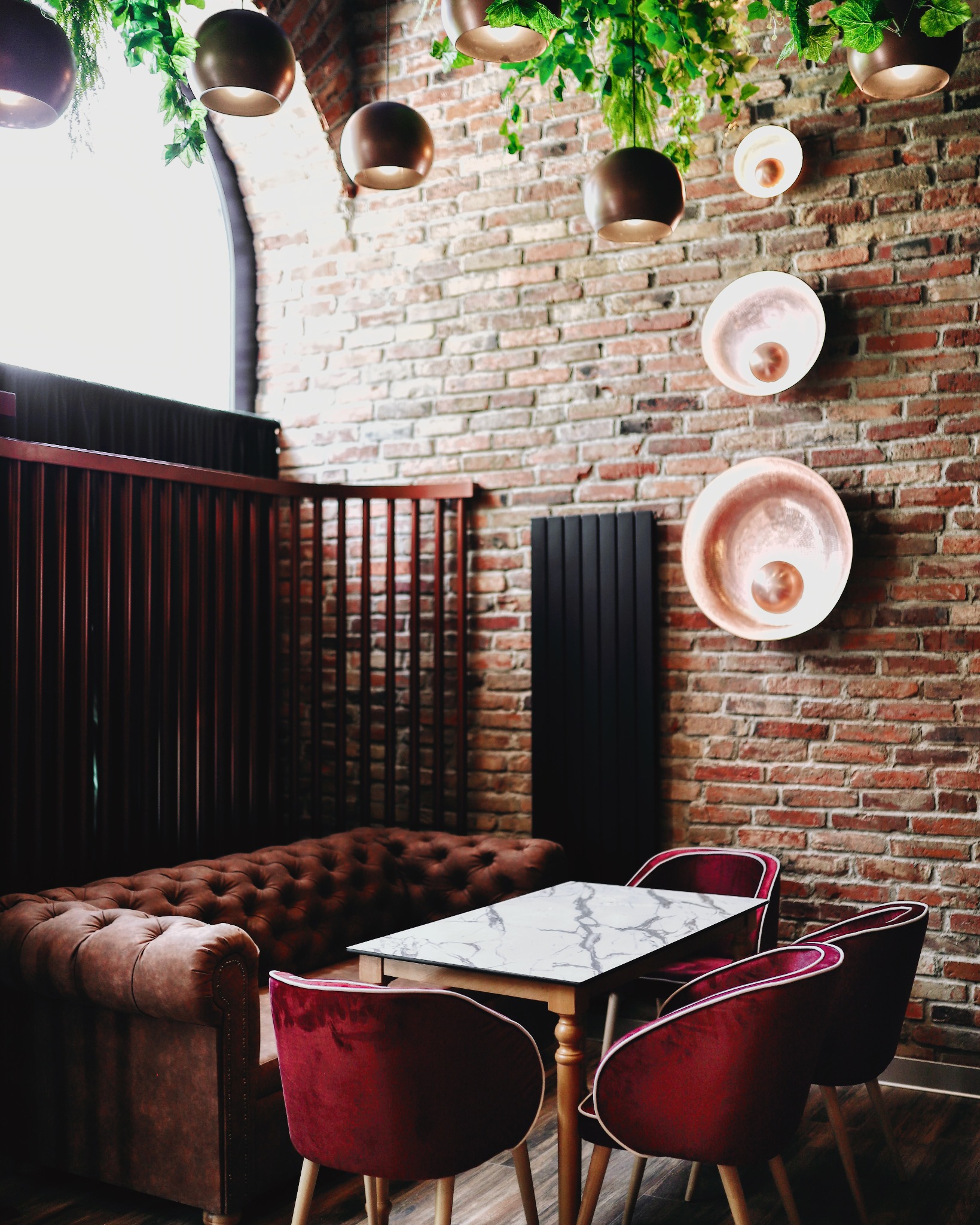 Stylowe wnętrze restauracji z ceglaną ścianą, skórzaną sofą Chesterfield, marmurowym stolikiem i krzesłami z bordowego weluru, oświetlone wiszącymi lampami i roślinami.