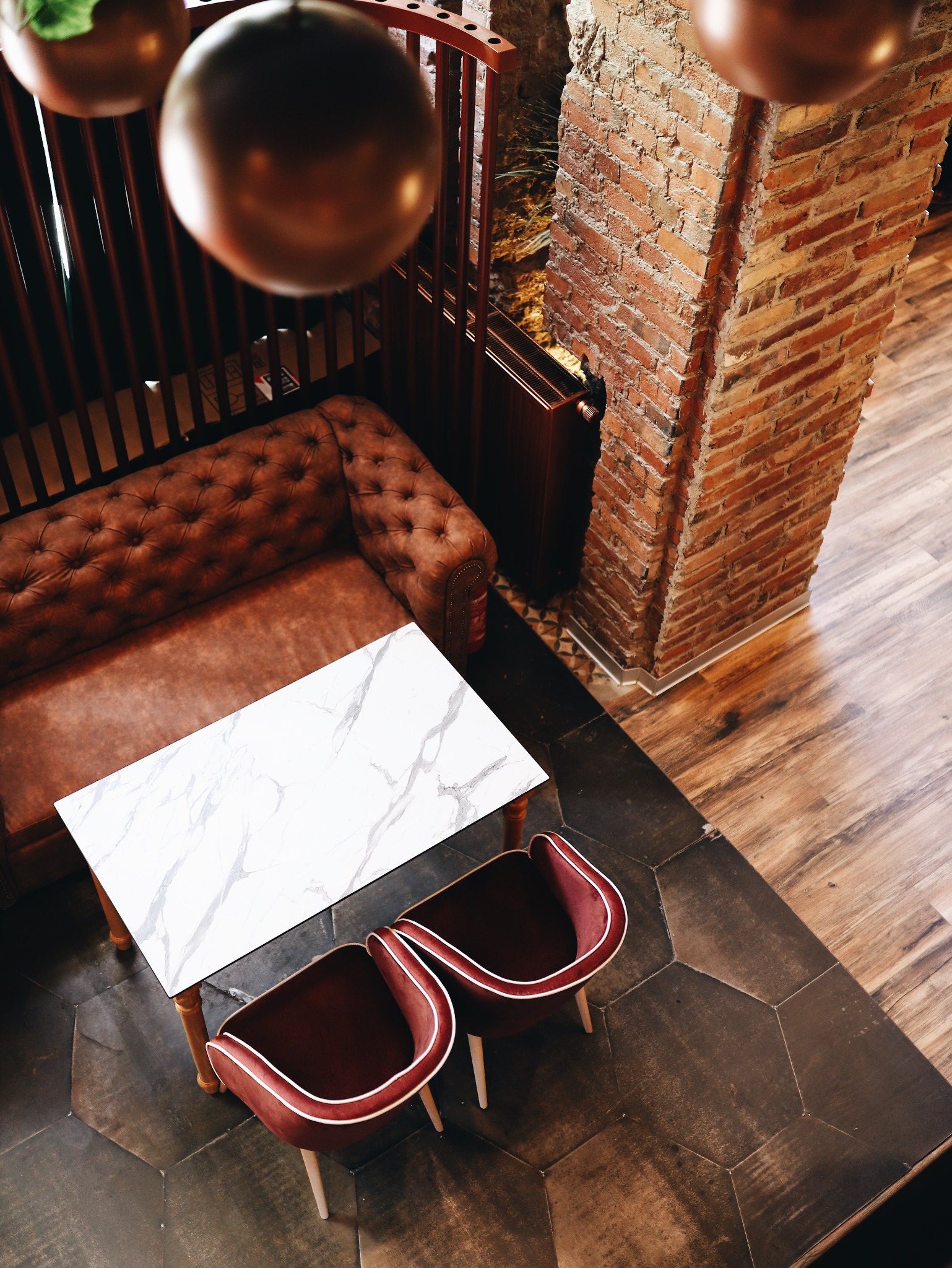 Widok z góry na eleganckie wnętrze z brązową skórzaną sofą typu chesterfield, marmurowym stołem, dwoma krzesłami z bordową tapicerką i ceglanym filarem, całość oświetlona ciepłym światłem wiszących...