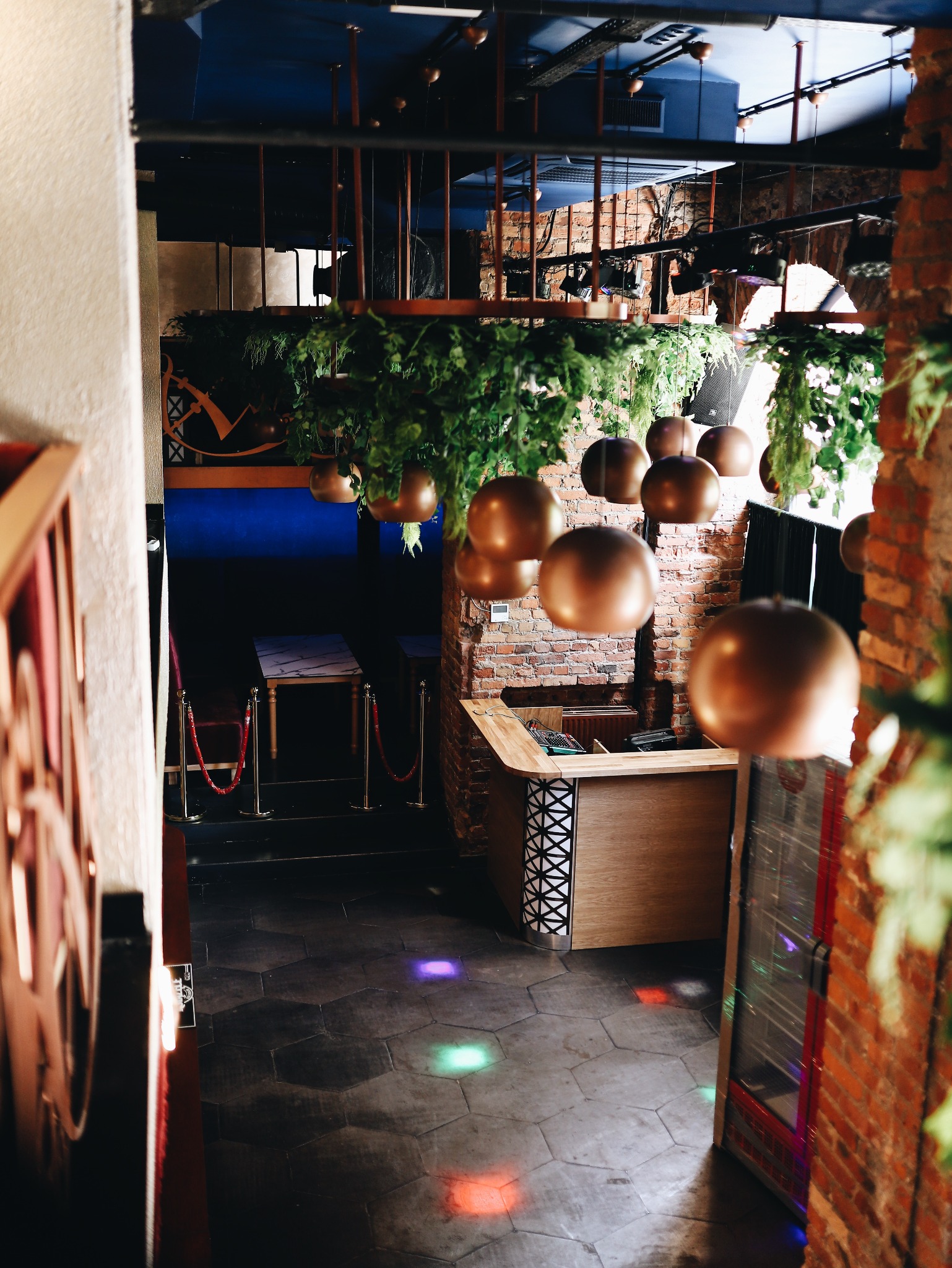 Wnętrze klubu z dekoracjami: wiszące rośliny, miedziane kule, ceglana ściana, parkiet z kolorowymi światłami, stoliki oddzielone czerwonymi sznurami.