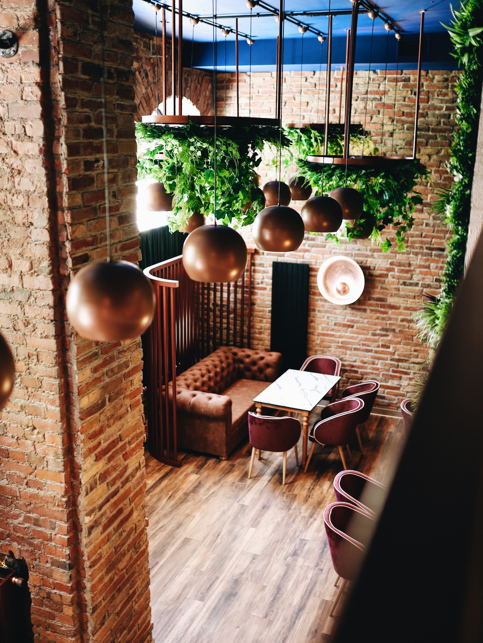 Wnętrze lokalu gastronomicznego z ceglanymi ścianami, drewnianą podłogą, pluszowymi krzesłami i sofą, marmurowym stolikiem oraz dekoracyjnymi lampami kulistymi i roślinami wiszącymi.