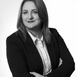 Portret kobiety w czarnym żakiecie i białej koszuli, z założonymi rękami i zegarkiem na nadgarstku, na jasnym tle w czerni i bieli.