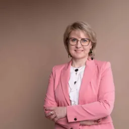 Portret uśmiechniętej kobiety w okularach i różowym żakiecie, skrzyżowane ręce, na beżowym tle.