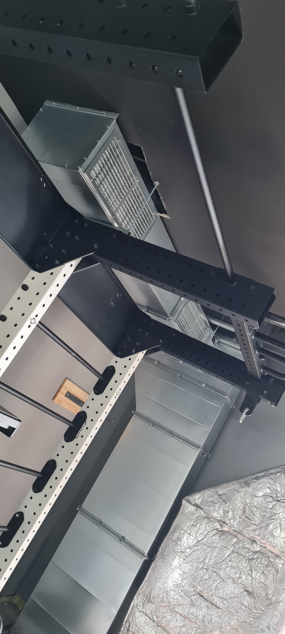 Widok z dołu na fragment systemu wentylacyjnego z metalowymi kanałami i kratkami wentylacyjnymi pod stropem, z elementami konstrukcji wsporczej w kolorze czarnym i białym.