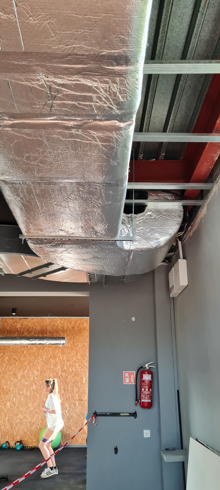 Widok na izolowany kanał wentylacyjny pod sufitem w sali ćwiczeń, z widocznym fragmentem instalacji elektrycznej i gaśnicą na ścianie. W tle ćwicząca osoba.
