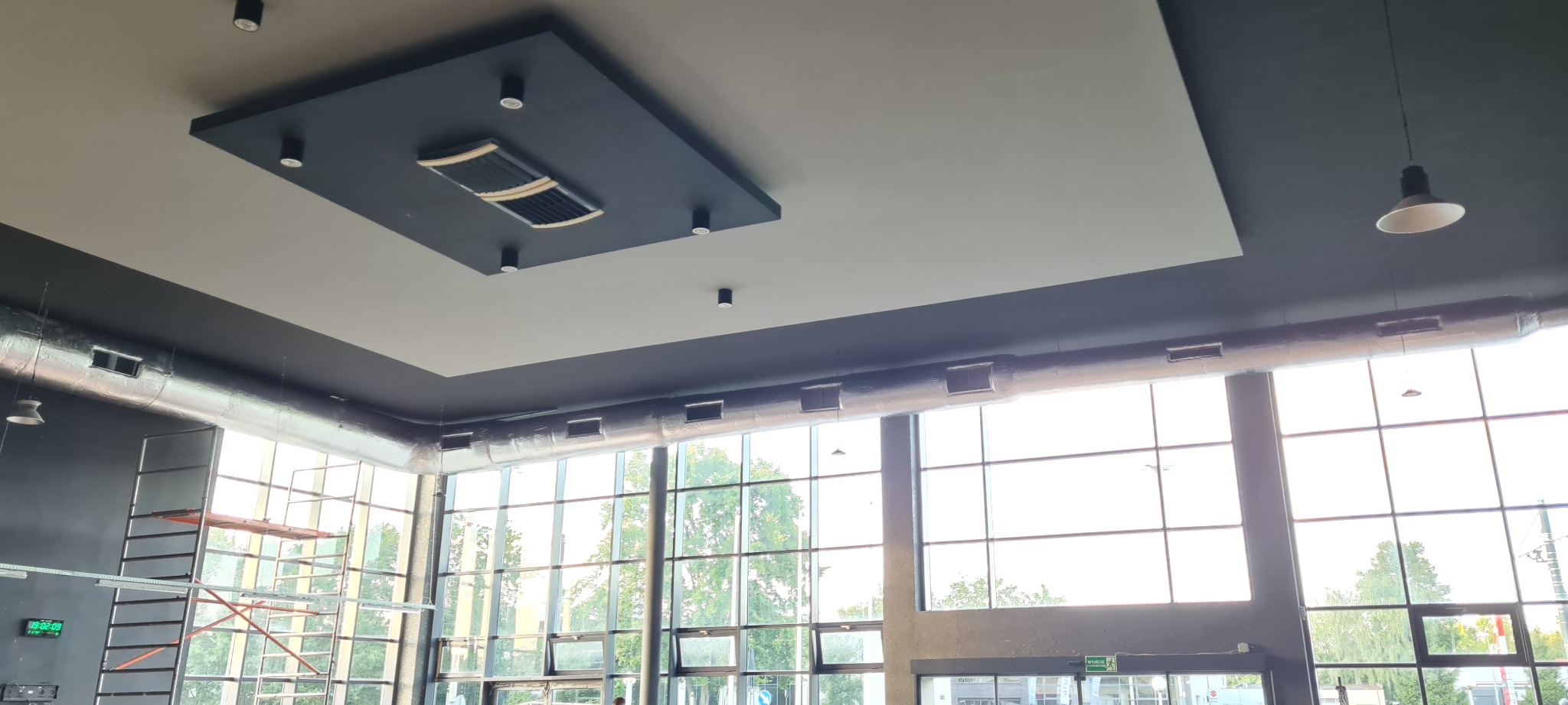Wnętrze hali z widoczną instalacją wentylacyjną pod sufitem, w tle duże okna i rusztowanie.