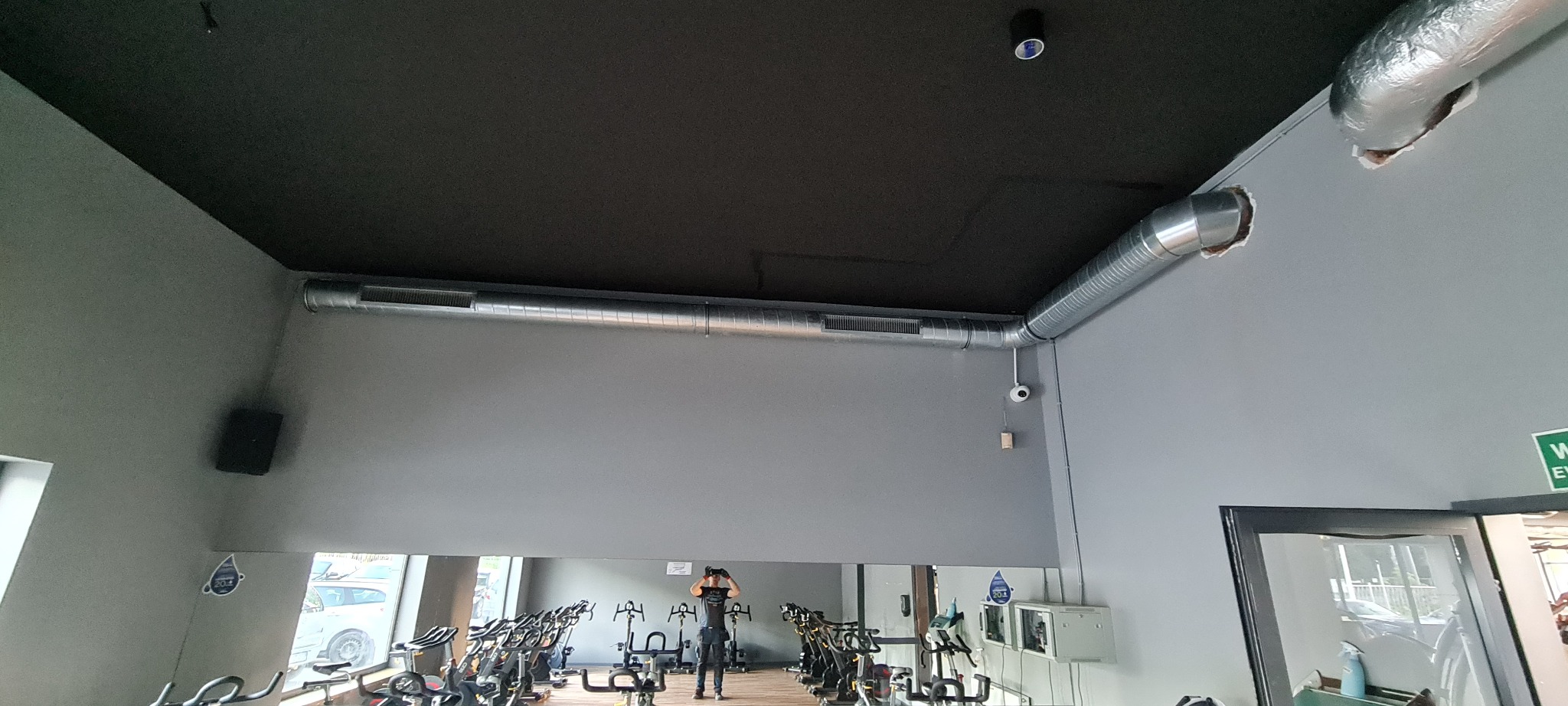 Szara sala fitness z zamontowaną pod sufitem srebrną, spiralną rurą wentylacyjną z widocznymi otworami wentylacyjnymi, czarnym sufitem i kamerą monitoringu na ścianie. W lustrze odbijają się rowery...