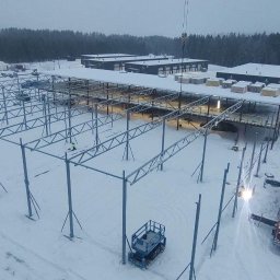 MKM Konstrukcje Stalowe - Montaż stalowej konstrukcji hali w zimowych warunkach, widoczny dźwig, podnośnik koszowy i pracownik na dachu istniejącej części hali. Śnieg pokrywa teren budowy i dachy sąsiednich budynków.