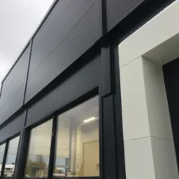 Zewnętrzny widok hali przemysłowej z czarną elewacją z płyt warstwowych, widoczne okna z odbiciami otoczenia i białe obramowanie wejścia.