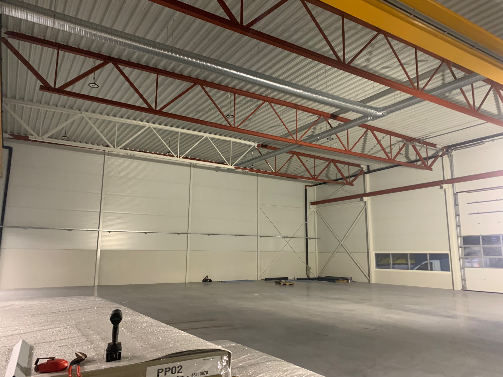 Wnętrze nowej hali produkcyjnej z widoczną konstrukcją stalową dachu pomalowaną na czerwono, systemem wentylacji pod sufitem i przygotowanymi do montażu płytami warstwowymi w dolnej części kadru.