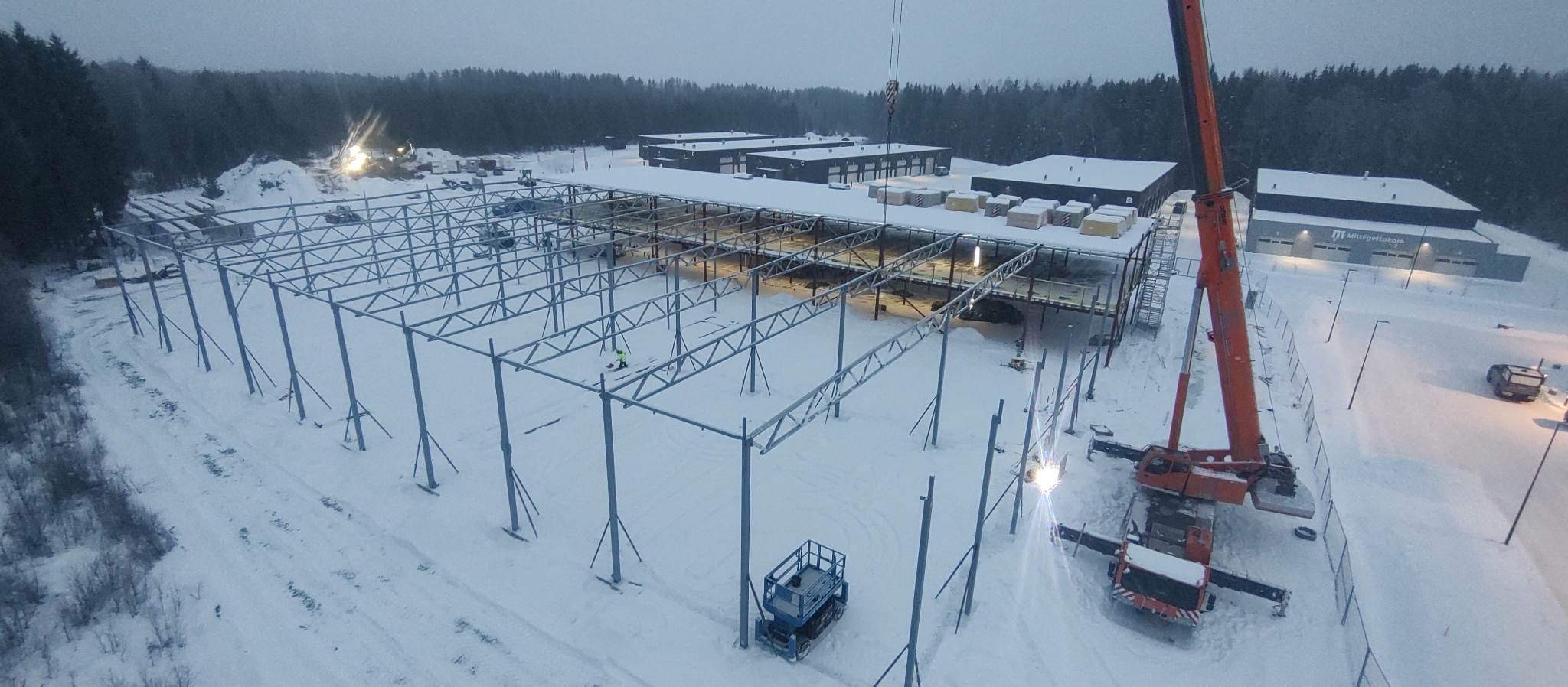 Montaż stalowej konstrukcji hali w zimowych warunkach, widoczny dźwig, podnośnik koszowy i pracownik na dachu istniejącej części hali. Śnieg pokrywa teren budowy i dachy sąsiednich budynków.
