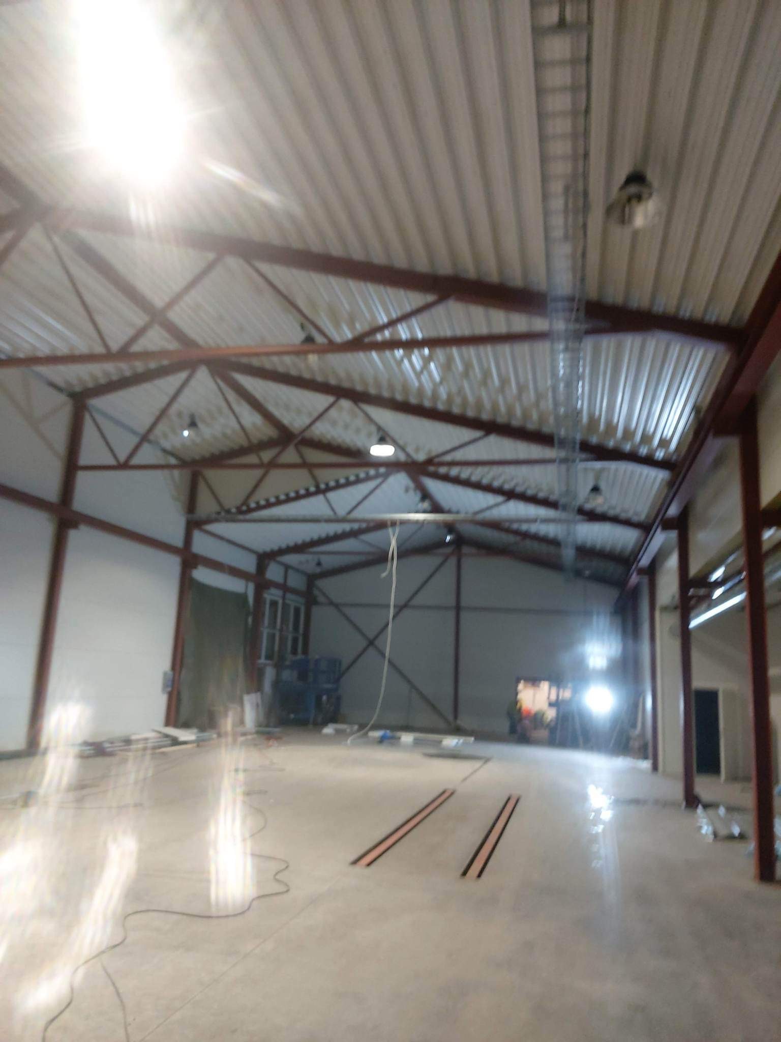 Wnętrze hali w trakcie budowy, widoczna stalowa konstrukcja dachu, oświetlenie oraz częściowo wykończone ściany. Na podłodze widoczne przewody i elementy montażowe.