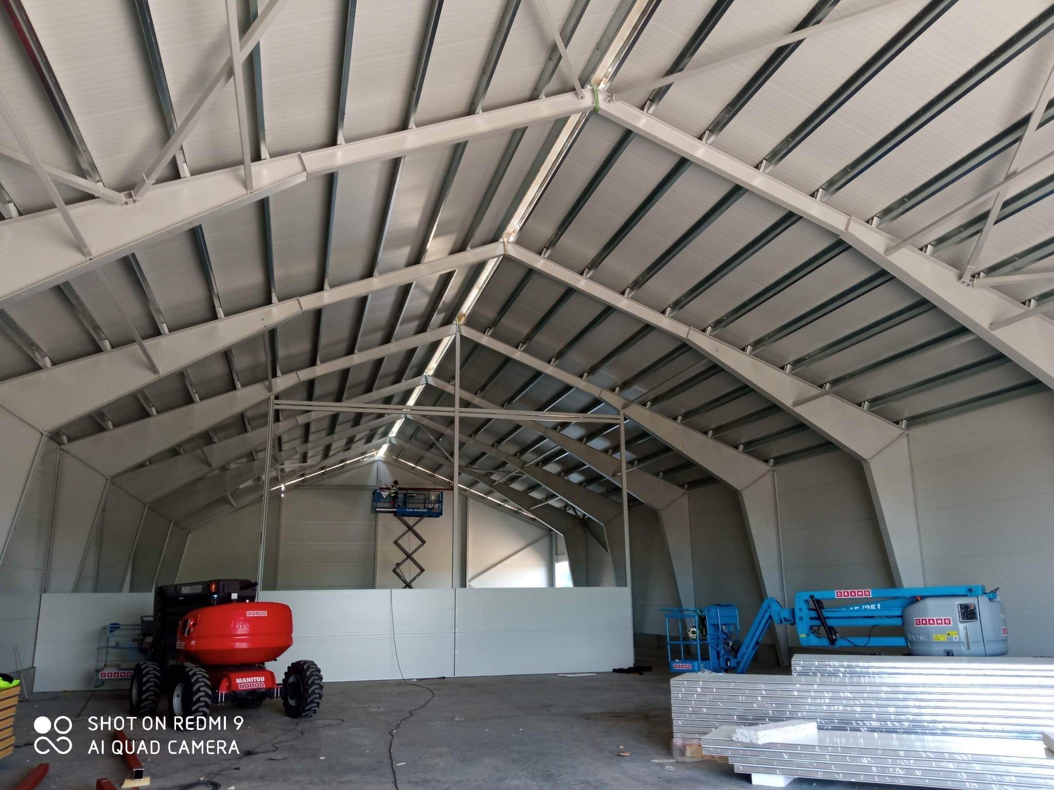 Wnętrze hali w trakcie budowy, widoczne stalowe elementy konstrukcyjne dachu, ściany z płyt warstwowych oraz podnośniki koszowe Manitou i Genie.