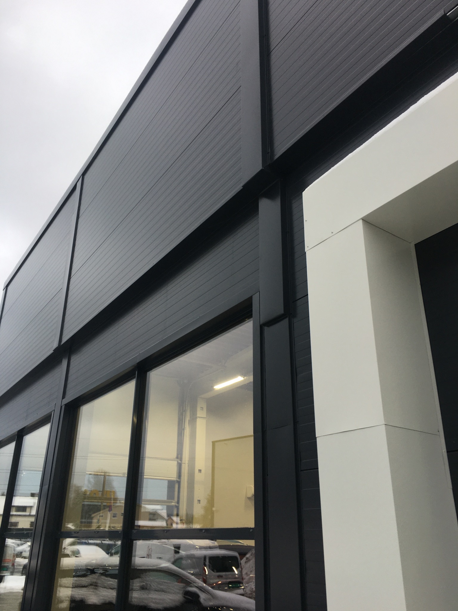 Zewnętrzny widok hali przemysłowej z czarną elewacją z płyt warstwowych, widoczne okna z odbiciami otoczenia i białe obramowanie wejścia.