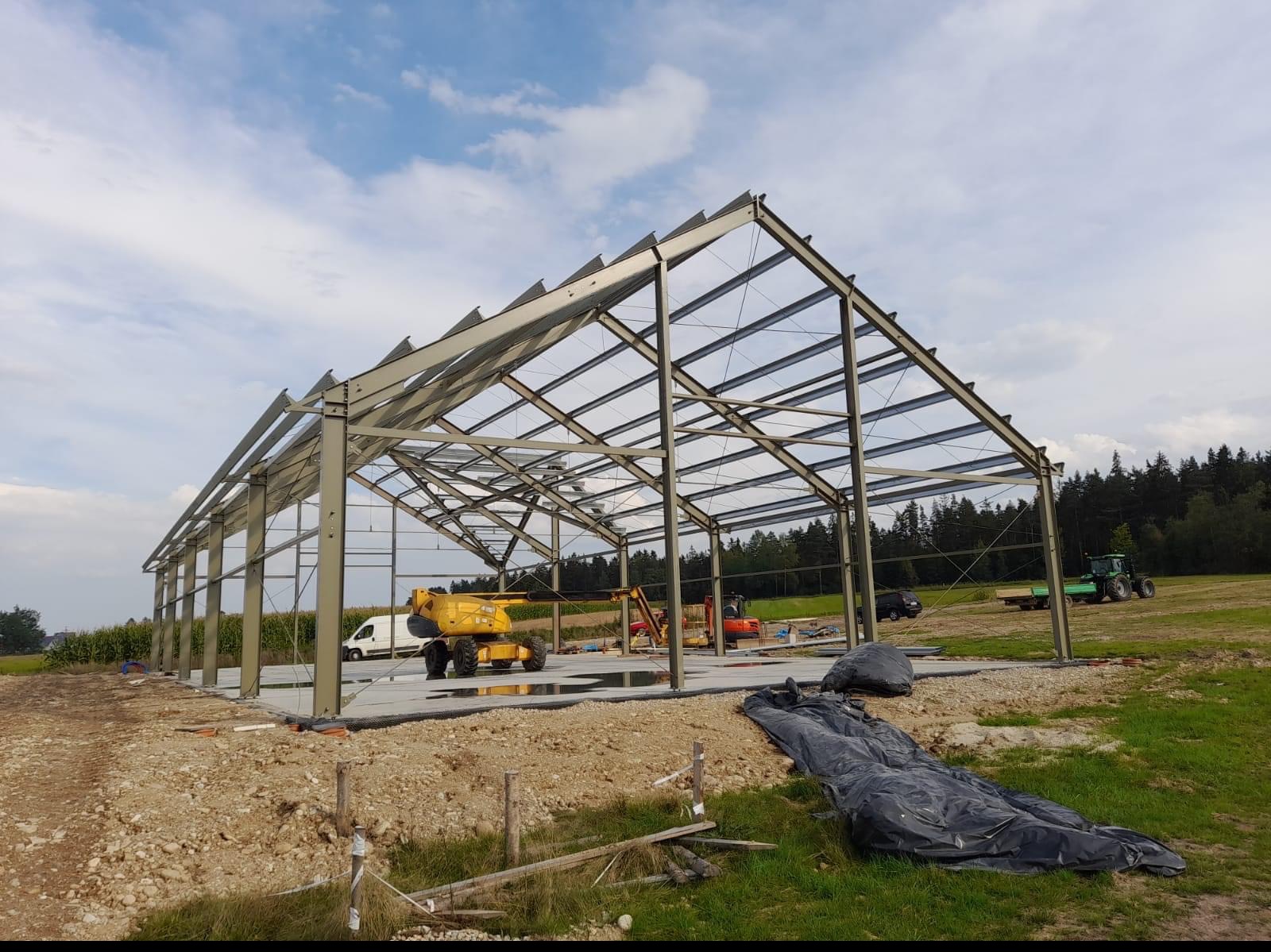 Stalowa konstrukcja szkieletowa hali na betonowej podstawie, z żółtym podnośnikiem koszowym i innymi maszynami budowlanymi w tle, na tle pola i lasu pod błękitnym niebem.