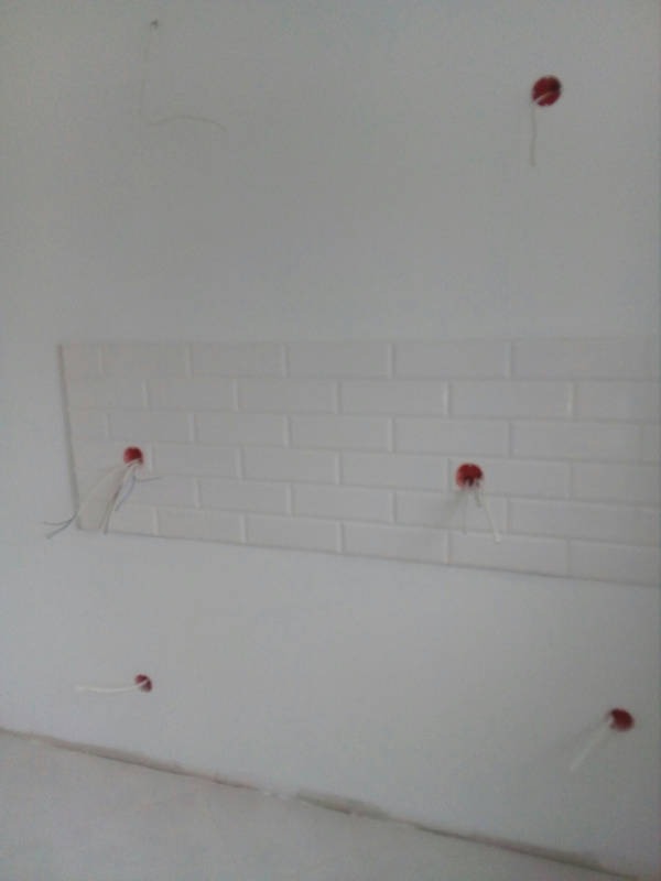 Ściana z białymi płytkami imitującymi cegłę, widoczne wystające kable elektryczne z czerwonymi osłonkami, etap prac wykończeniowych.