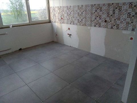 Remontowana kuchnia: szare płytki podłogowe ułożone z krzyżykami dystansowymi, częściowo położone dekoracyjne płytki ścienne, widoczne instalacje hydrauliczne i elektryczne przed wykończeniem.