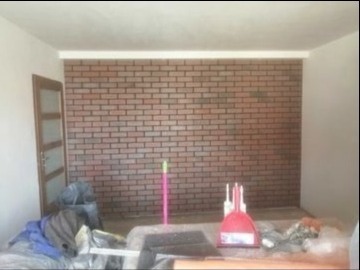 Wnętrze pokoju po częściowym remoncie, ściana wykończona panelami imitującymi czerwoną cegłę, widoczne narzędzia malarskie i worki z materiałami budowlanymi, sufit pomalowany na biało, widoczny...