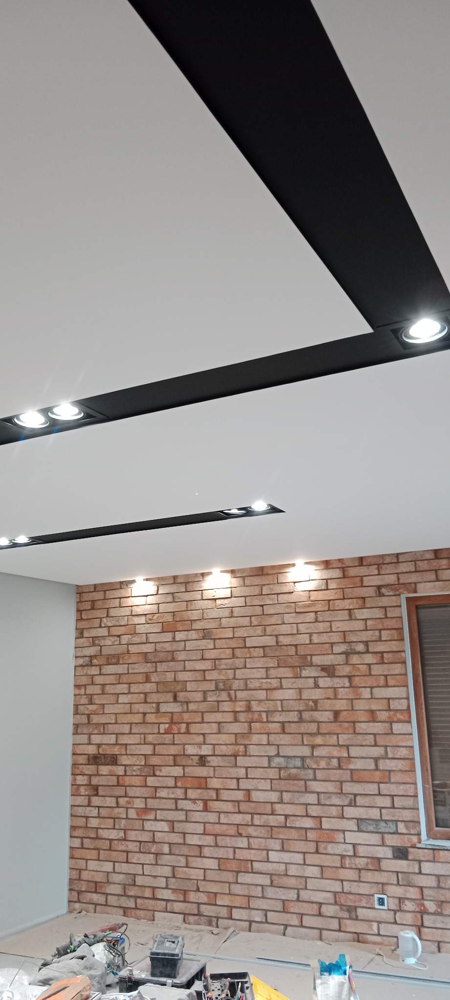 Ściana z cegły dekoracyjnej podświetlona punktowymi lampami sufitowymi w nowoczesnym wnętrzu, widoczne narzędzia pracy na podłodze.