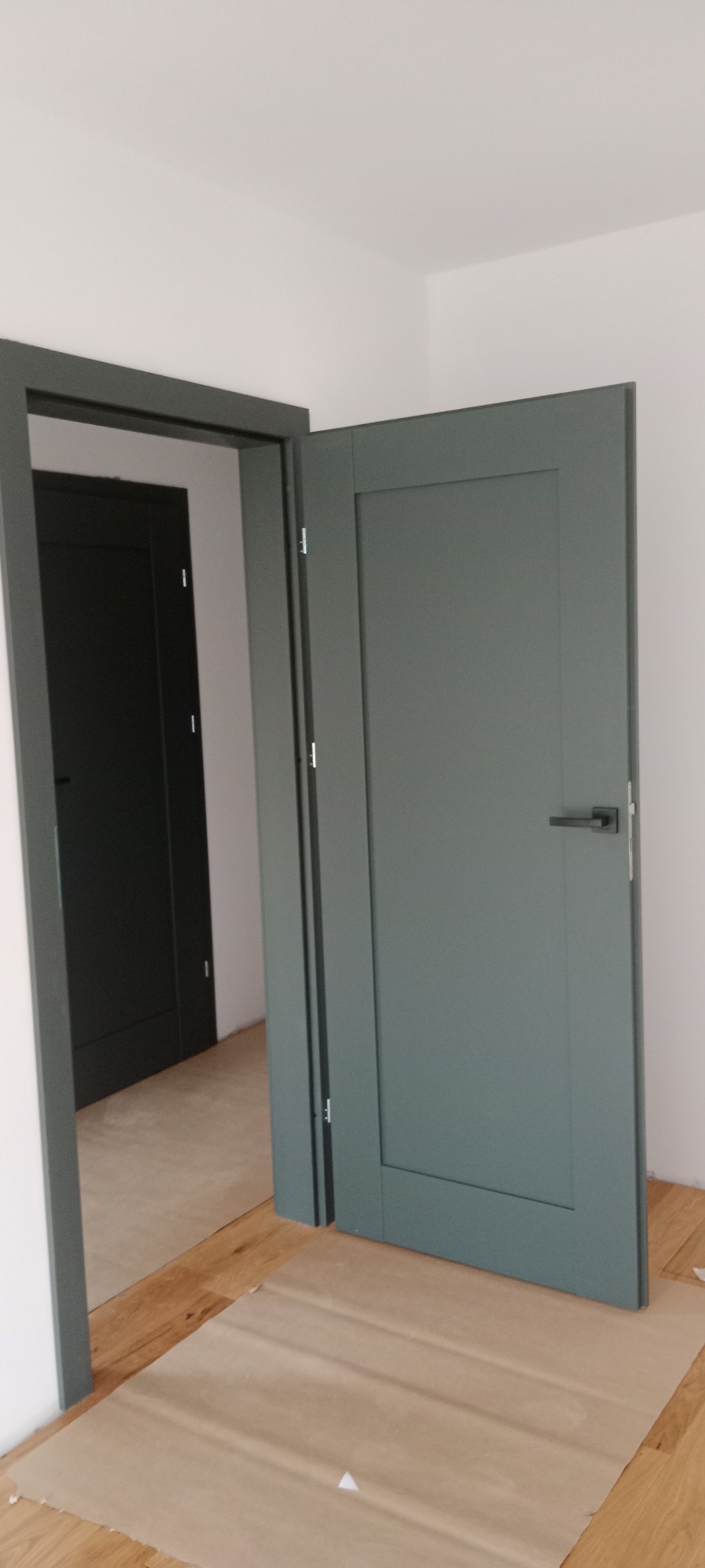 Szare, panelowe drzwi wewnętrzne z czarną klamką, częściowo otwarte, widoczne zabezpieczenie podłogi kartonem, w tle kolejne drzwi w ościeżnicy.