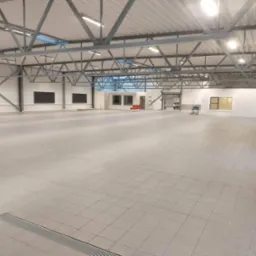 Wnętrze nowo wybudowanej hali z widoczną konstrukcją stalową dachu, szarą posadzką z płytek i białymi ścianami z oknami. W oddali widoczne krzesła i drabina.