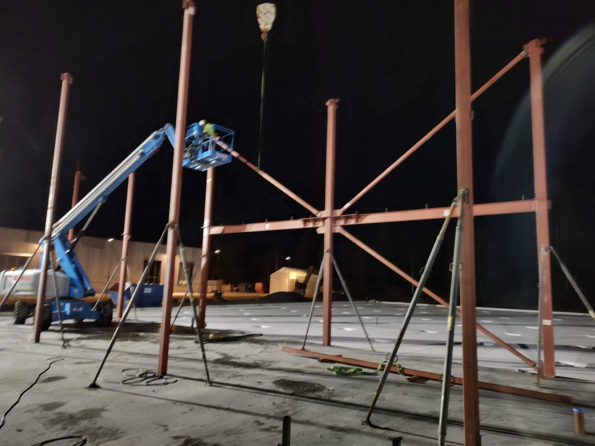 Konstrukcja stalowa hali w trakcie montażu nocą, widoczny podnośnik koszowy z pracownikiem, elementy konstrukcyjne podtrzymywane przez podpory tymczasowe.