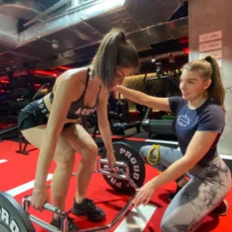 Kobieta wykonuje martwy ciąg z heksagonalnym gryfem pod nadzorem drugiej kobiety w siłowni z czerwono-czarną podłogą i widocznymi instalacjami pod sufitem.