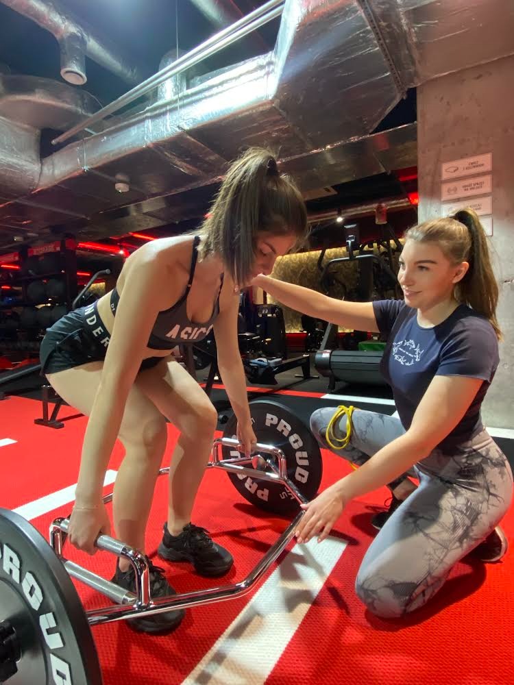 Kobieta wykonuje martwy ciąg z heksagonalnym gryfem pod nadzorem drugiej kobiety w siłowni z czerwono-czarną podłogą i widocznymi instalacjami pod sufitem.