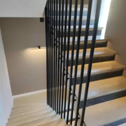 Nowoczesne schody z jasnego drewna z czarną, minimalistyczną balustradą. Widok z góry, oświetlenie ledowe pod stopniami tworzy subtelny efekt.