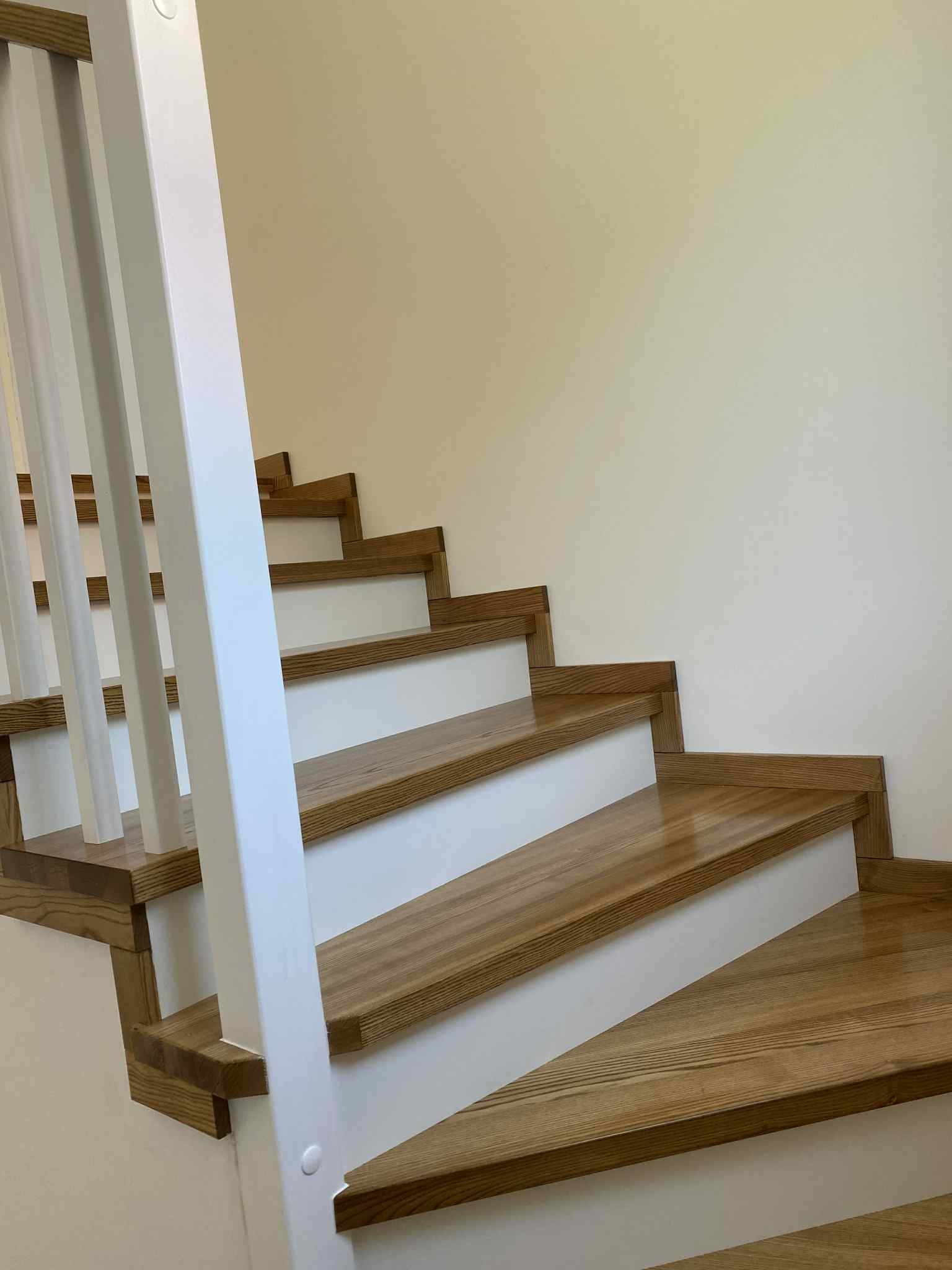 Jasne, drewniane schody z białymi podstopnicami i białą balustradą, widziane z dołu, z wyraźnym usłojeniem drewna i gładką powierzchnią stopni.
