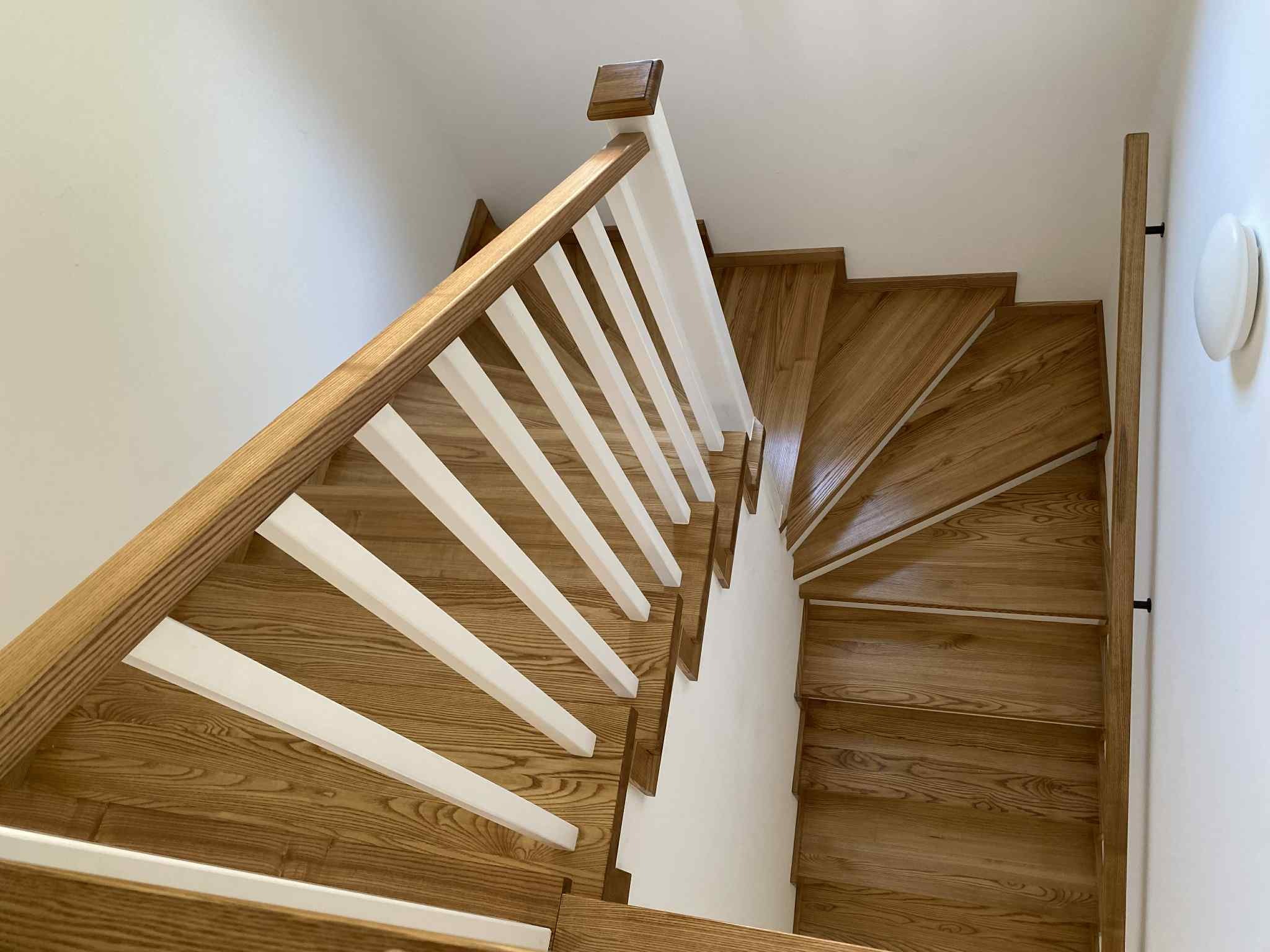 Kątowe ujęcie z góry na drewniane schody z balustradą. Jasne drewno stopni kontrastuje z bielą ściany i balustrady. Widoczny fragment oświetlenia na ścianie.