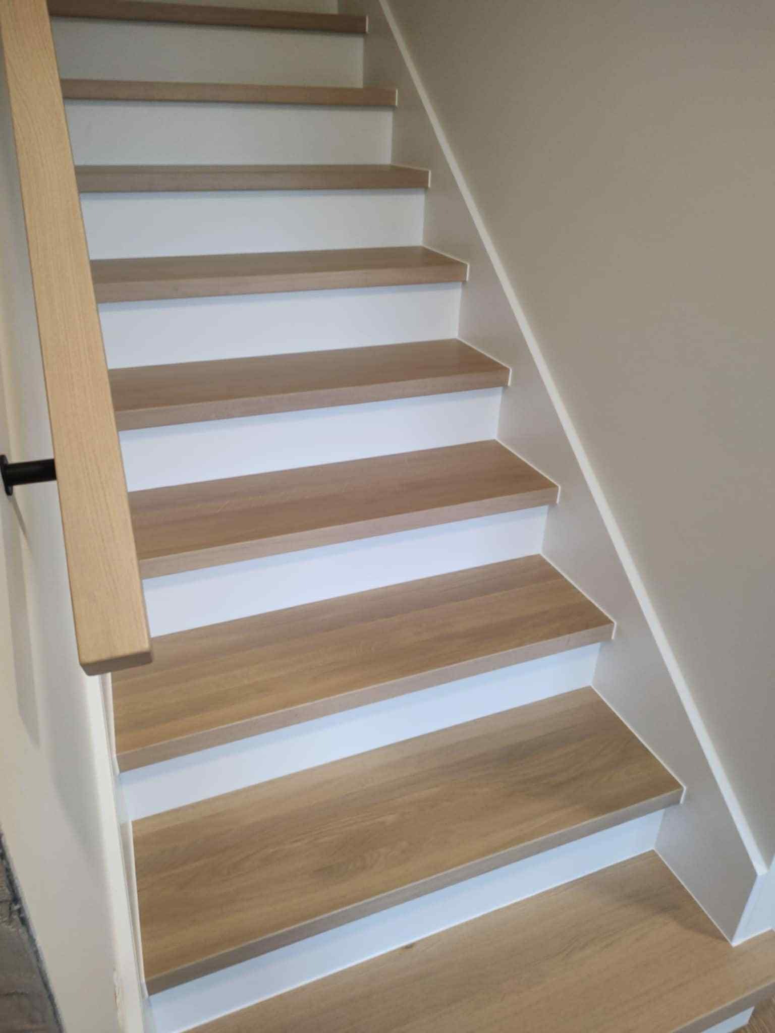Jasne schody z drewnianymi stopniami i białą podstopnicą, widok z dołu. Prosta, nowoczesna balustrada z jasnego drewna z czarnymi mocowaniami.