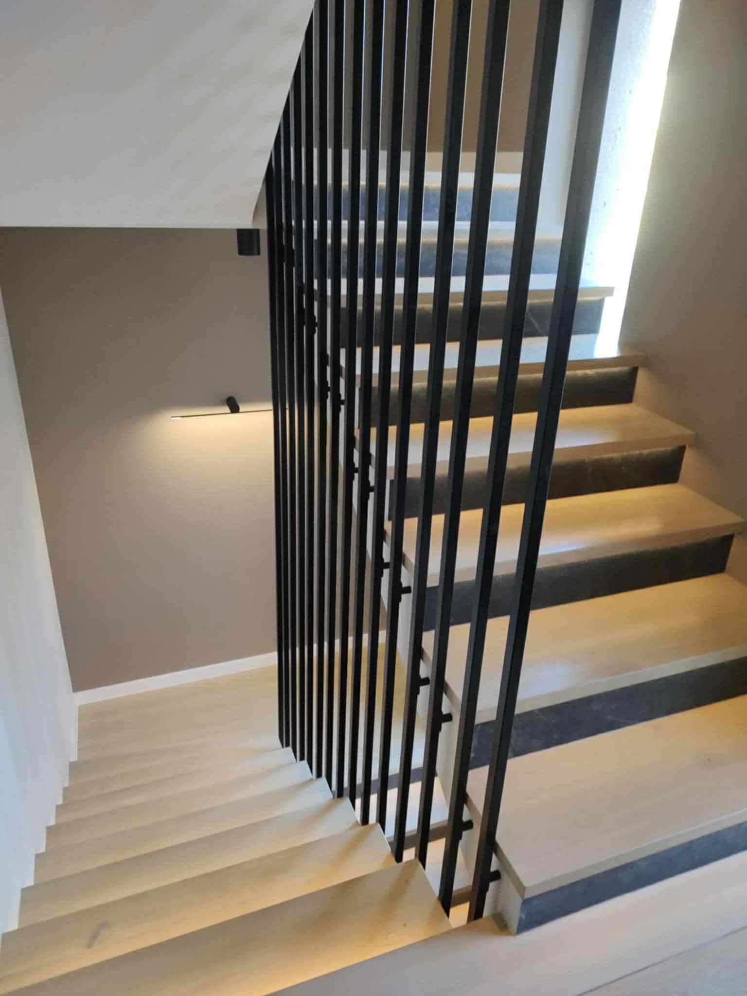 Nowoczesne schody z jasnego drewna z czarną, minimalistyczną balustradą. Widok z góry, oświetlenie ledowe pod stopniami tworzy subtelny efekt.