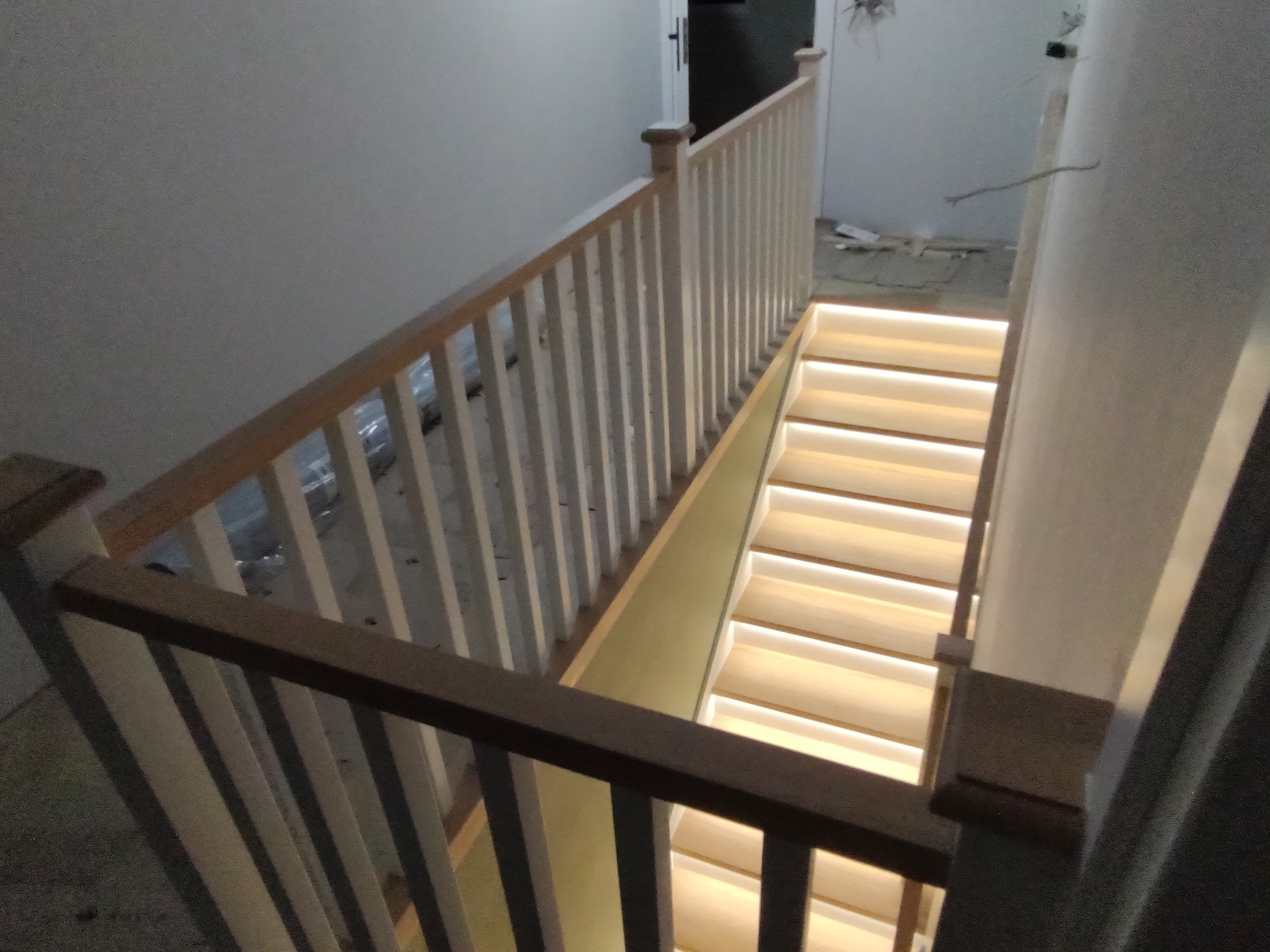 Widok z góry na jasne schody z podświetleniem LED i drewnianą, białą balustradą w trakcie montażu wewnątrz budynku. Surowy stan wykończenia wnętrza.