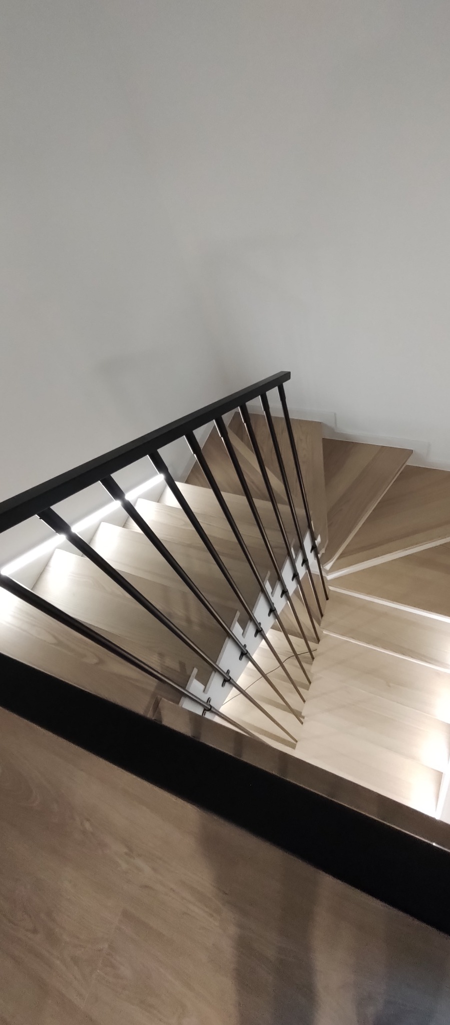 Spiralne schody z jasnego drewna, zabezpieczone nowoczesną, czarną balustradą z metalowymi prętami, widoczne z góry w minimalistycznym wnętrzu.