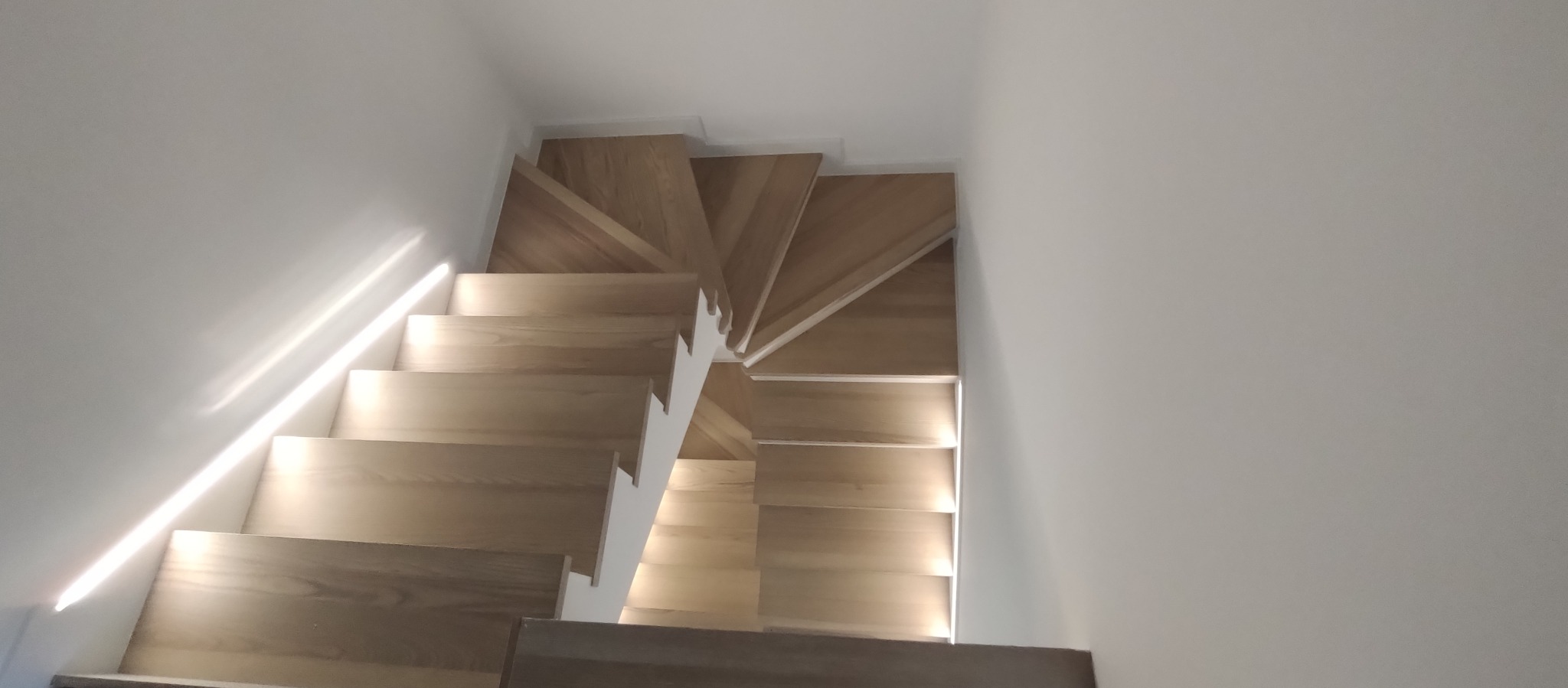 Kątowe ujęcie z góry na nowoczesne, drewniane schody z podświetleniem LED, w minimalistycznym wnętrzu z białymi ścianami. Widoczny fragment podłogi na dole.