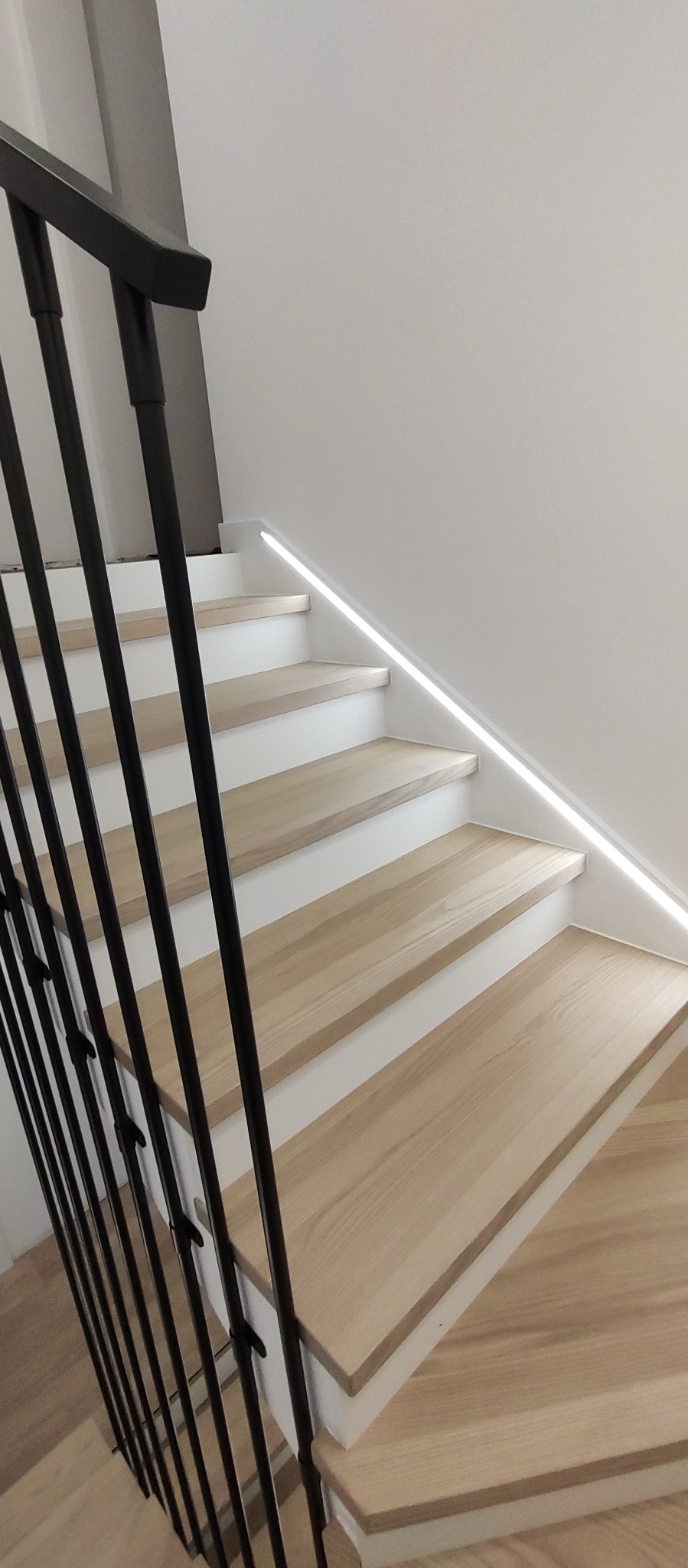 Jasne stopnie z oświetleniem LED wzdłuż ściany i czarna, metalowa balustrada. Minimalistyczny design schodów.