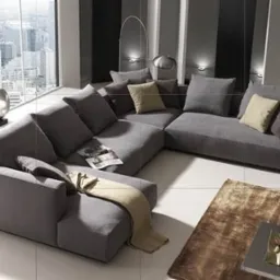 Szary, modułowy narożnik z poduszkami w salonie z widokiem na miasto, dywan w odcieniach brązu i stolik kawowy z dekoracjami.