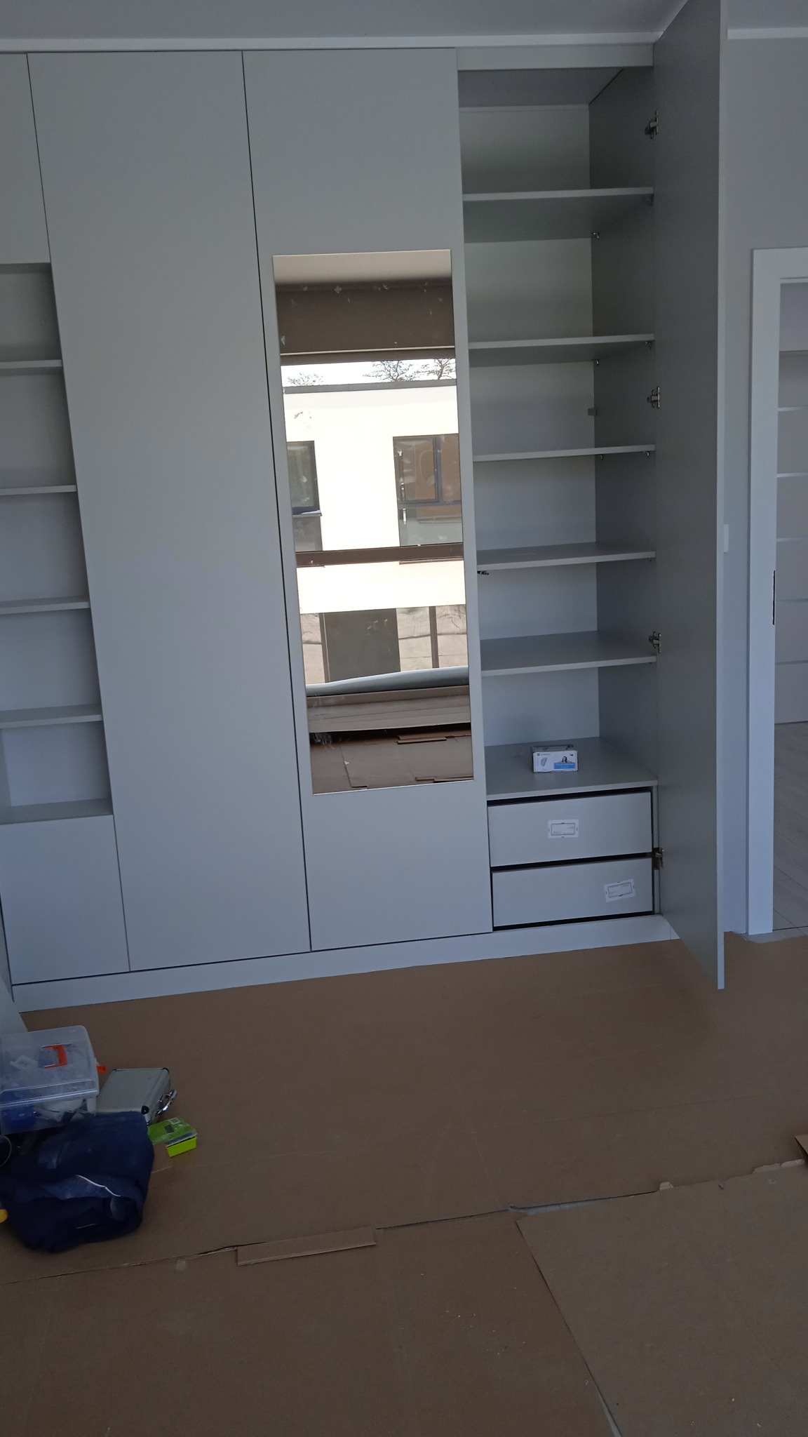 Biała szafa z lustrem w trakcie montażu, widoczne otwarte drzwi z półkami i szufladami, podłoga zabezpieczona kartonem, narzędzia w tle.