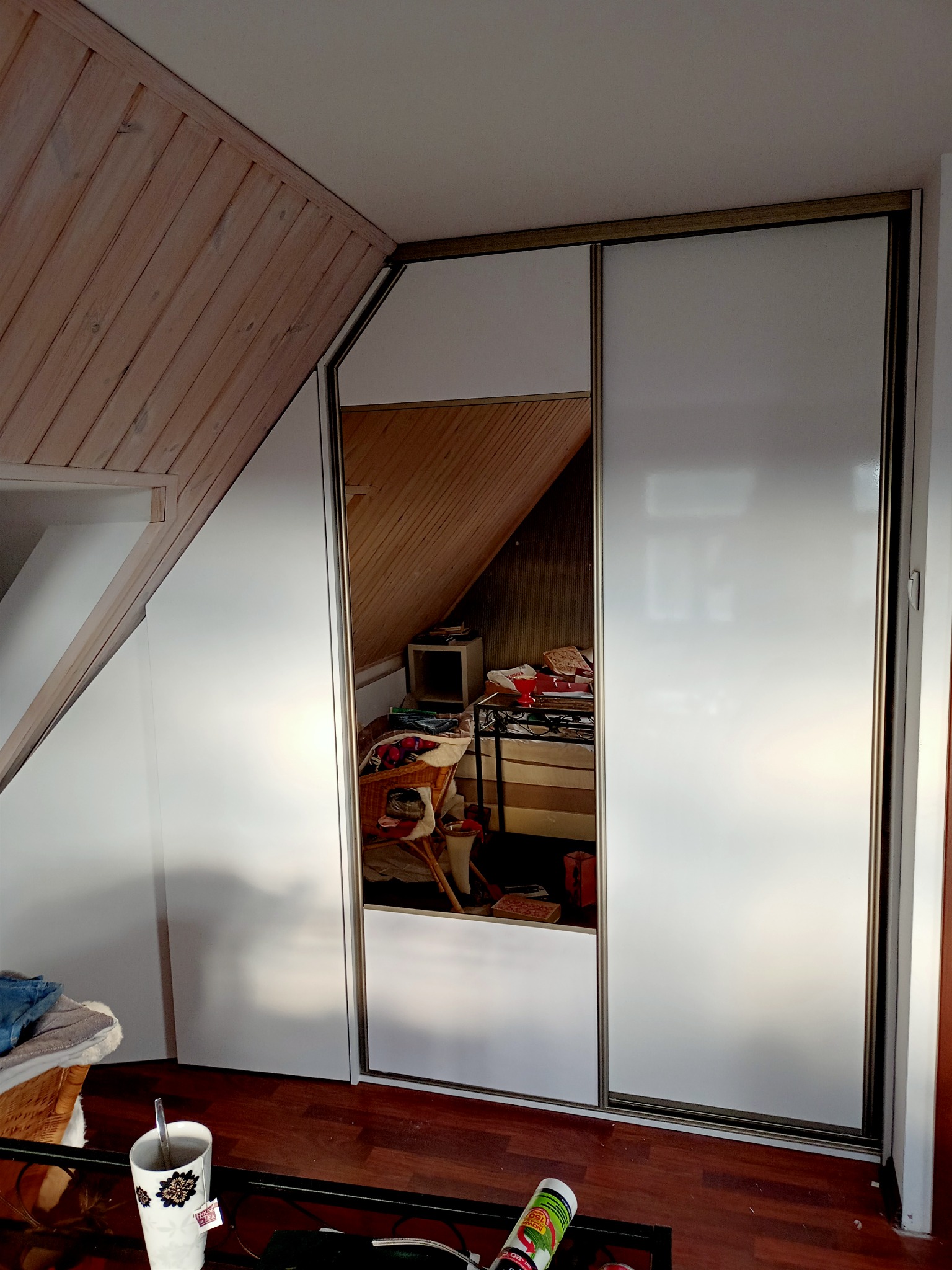 Biała szafa przesuwna z lustrem w skosie poddasza, widoczne elementy wyposażenia wnętrza w odbiciu.