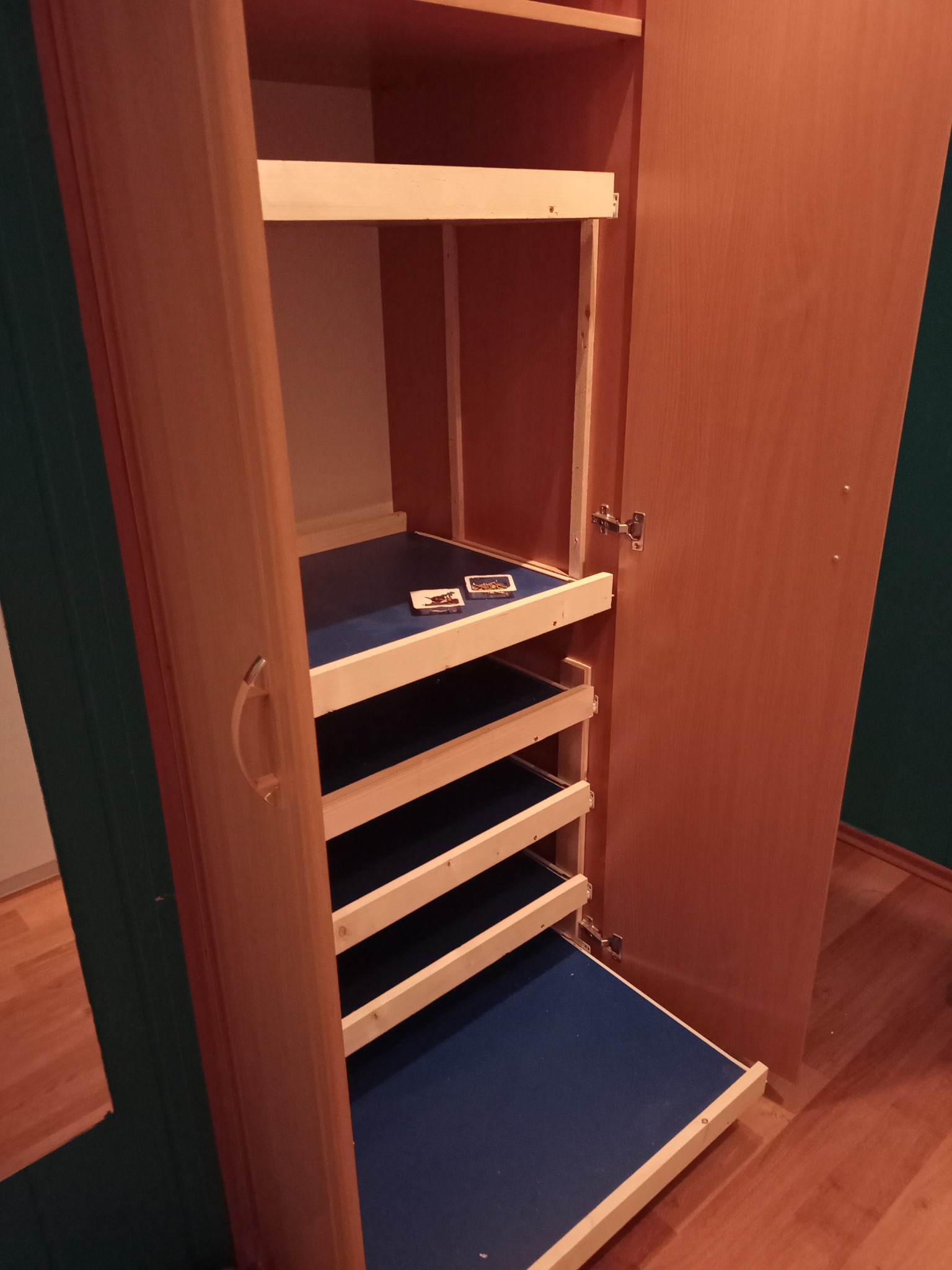 Otwarta szafa z jasnego drewna z wysuwanymi półkami w kolorze granatowym. Na jednej z półek leżą dwie karty.