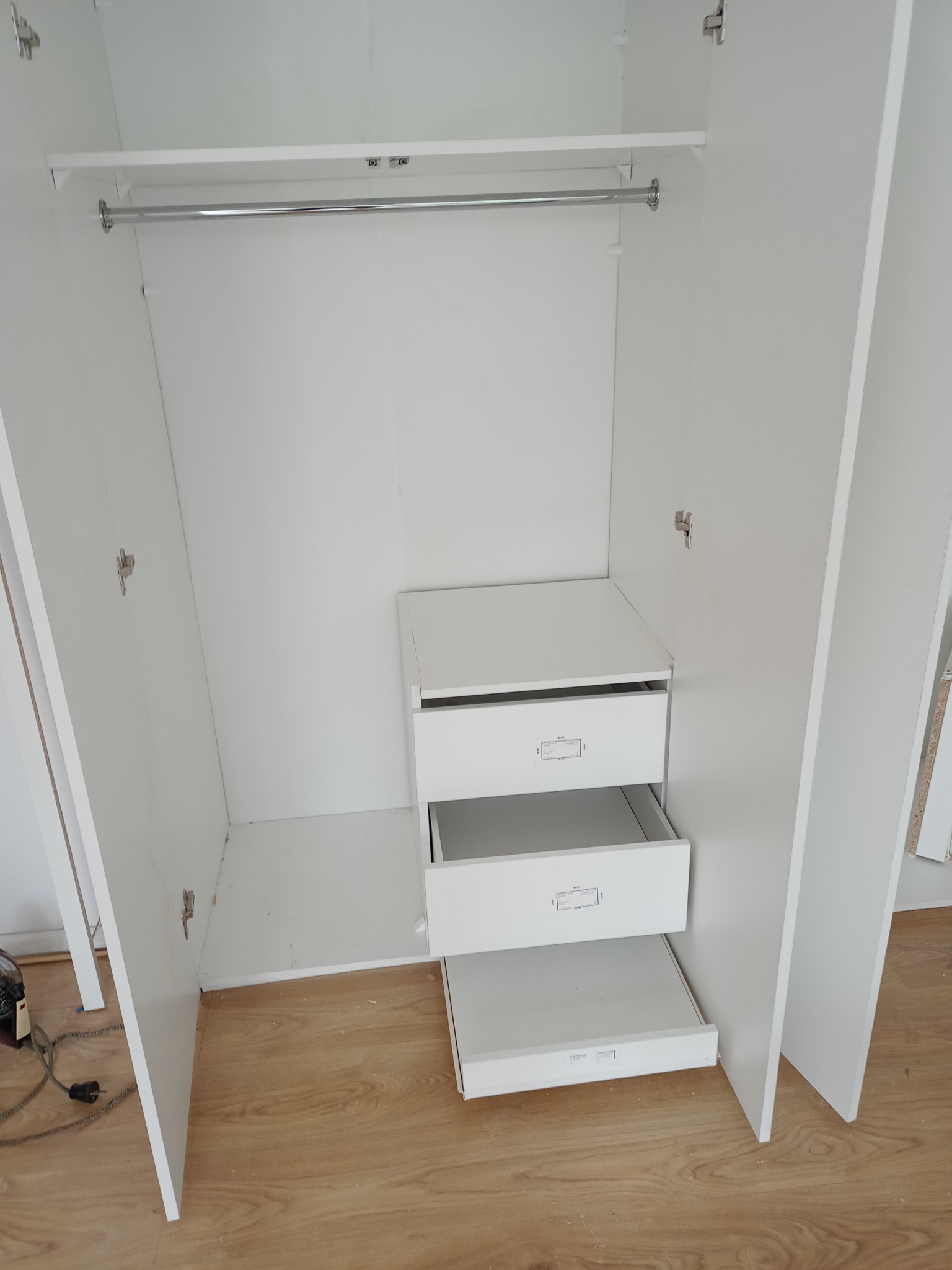 Wnętrze białej szafy z drążkiem na ubrania i trzema szufladami, z których jedna jest wysunięta, widok z góry na drewnianą podłogę.
