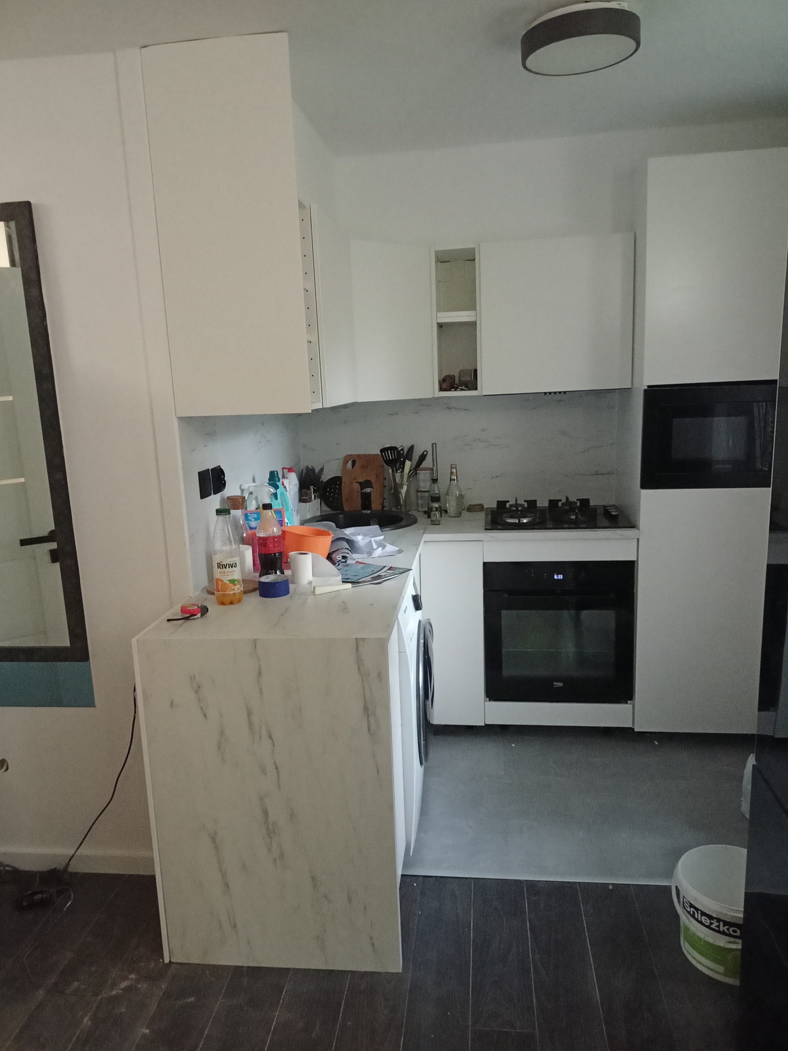 Wnętrze kuchni w trakcie montażu, białe szafki z marmurkowym blatem, widoczne narzędzia i materiały budowlane, ściana częściowo wykończona.