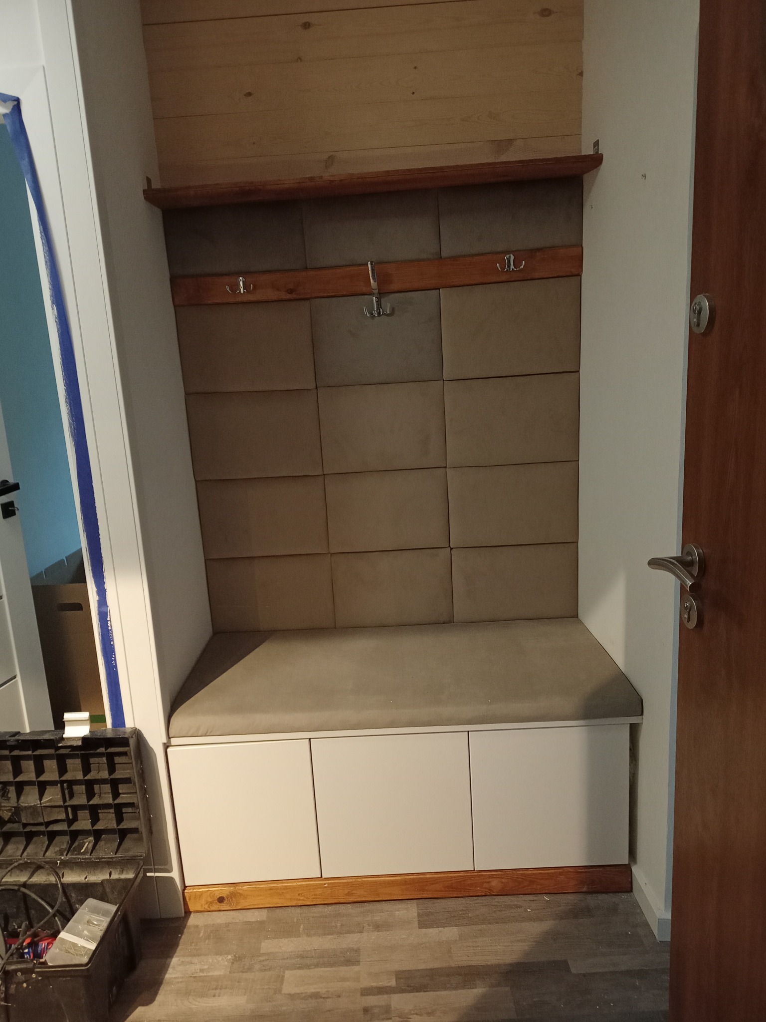 Wnęka w przedpokoju z siedziskiem i panelem tapicerowanym na ścianie, drewniana półka i wieszaki, widoczna skrzynka z narzędziami.