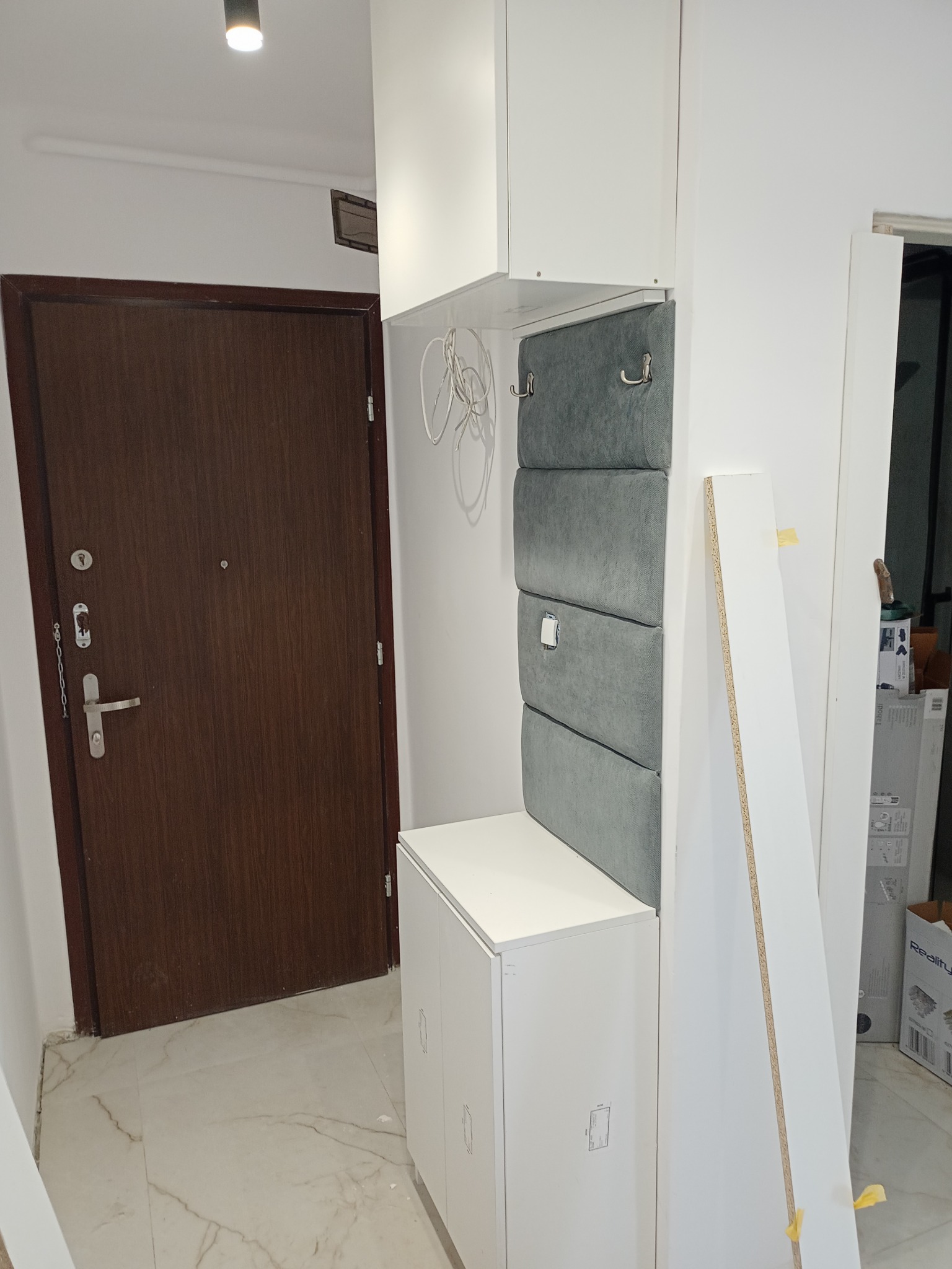 Przedpokój z zabudową na wymiar: biała szafka z siedziskiem obitym szarą tapicerką, wieszaki, brązowe drzwi wejściowe, marmurowa podłoga, elementy montażowe w tle.